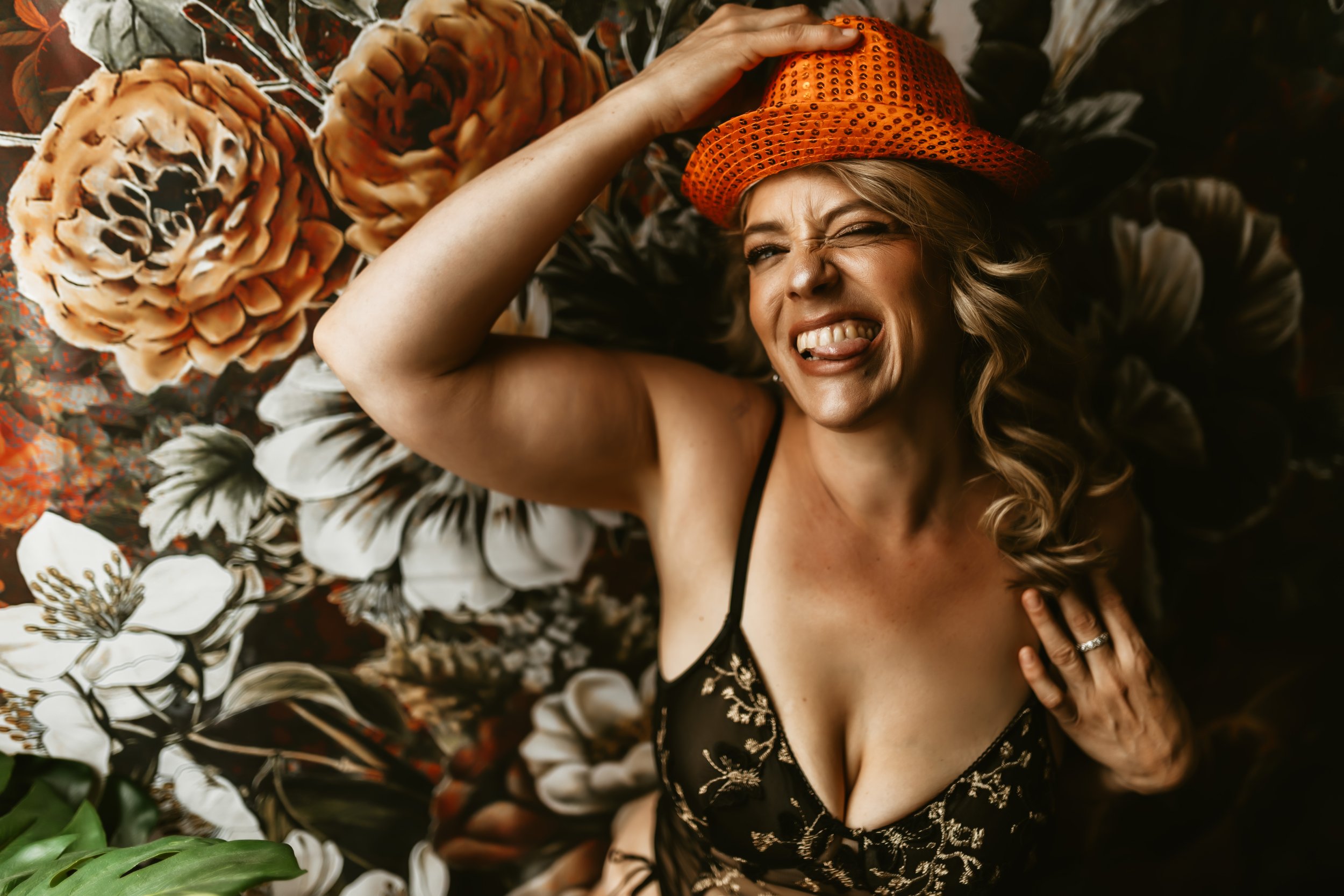 A woman lying on her back on a bed with a floral pattern, wearing a black dress with gold embroidery, an orange bucket hat, and making a playful face with her tongue out, eyes closed, and hand on the hat.