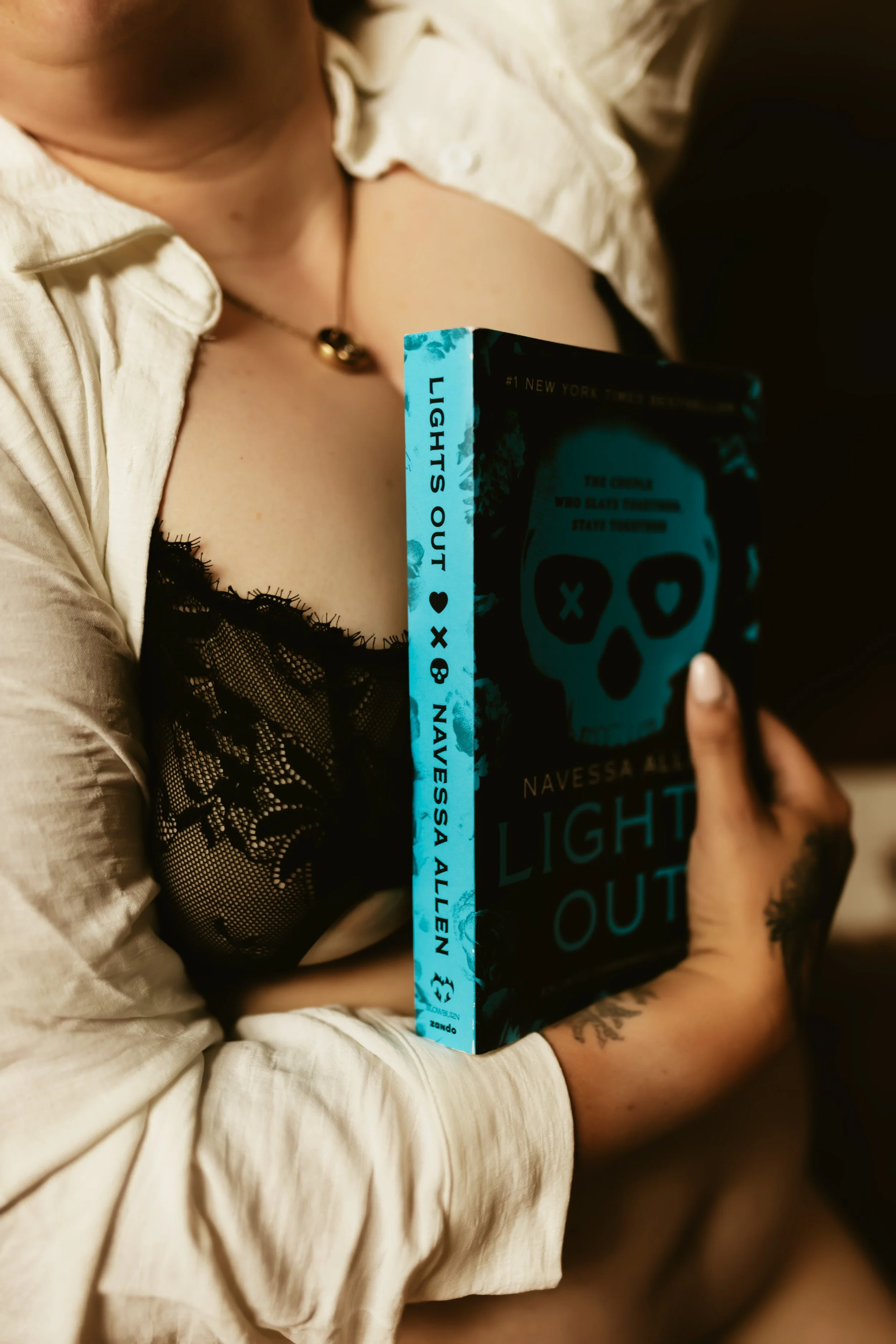 A person wearing a cream-colored shirt with lace detail on the sleeve, holding a copy of the book "Light Out" by Navessa Allen, with visible tattoos on their wrist and hand.