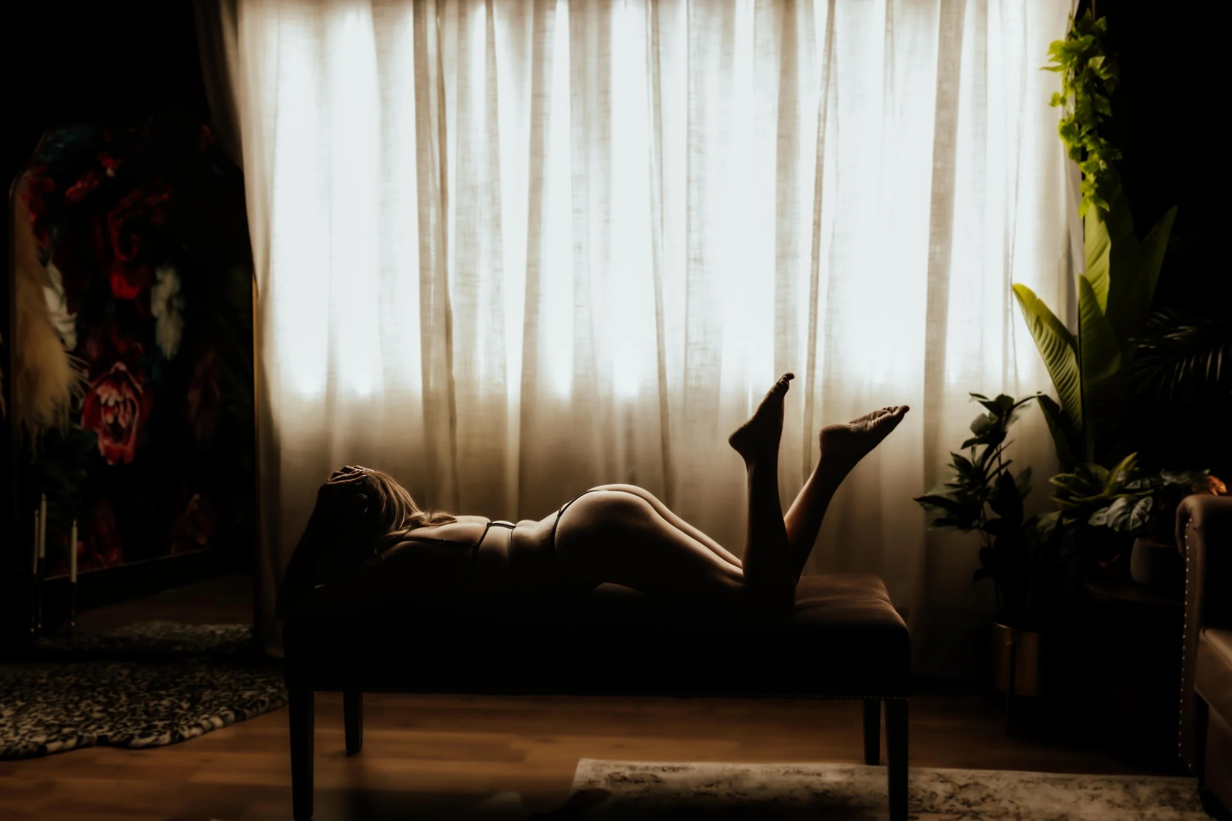 Person lying on bed with legs raised toward a window with curtains, surrounded by plants in a cozy room.