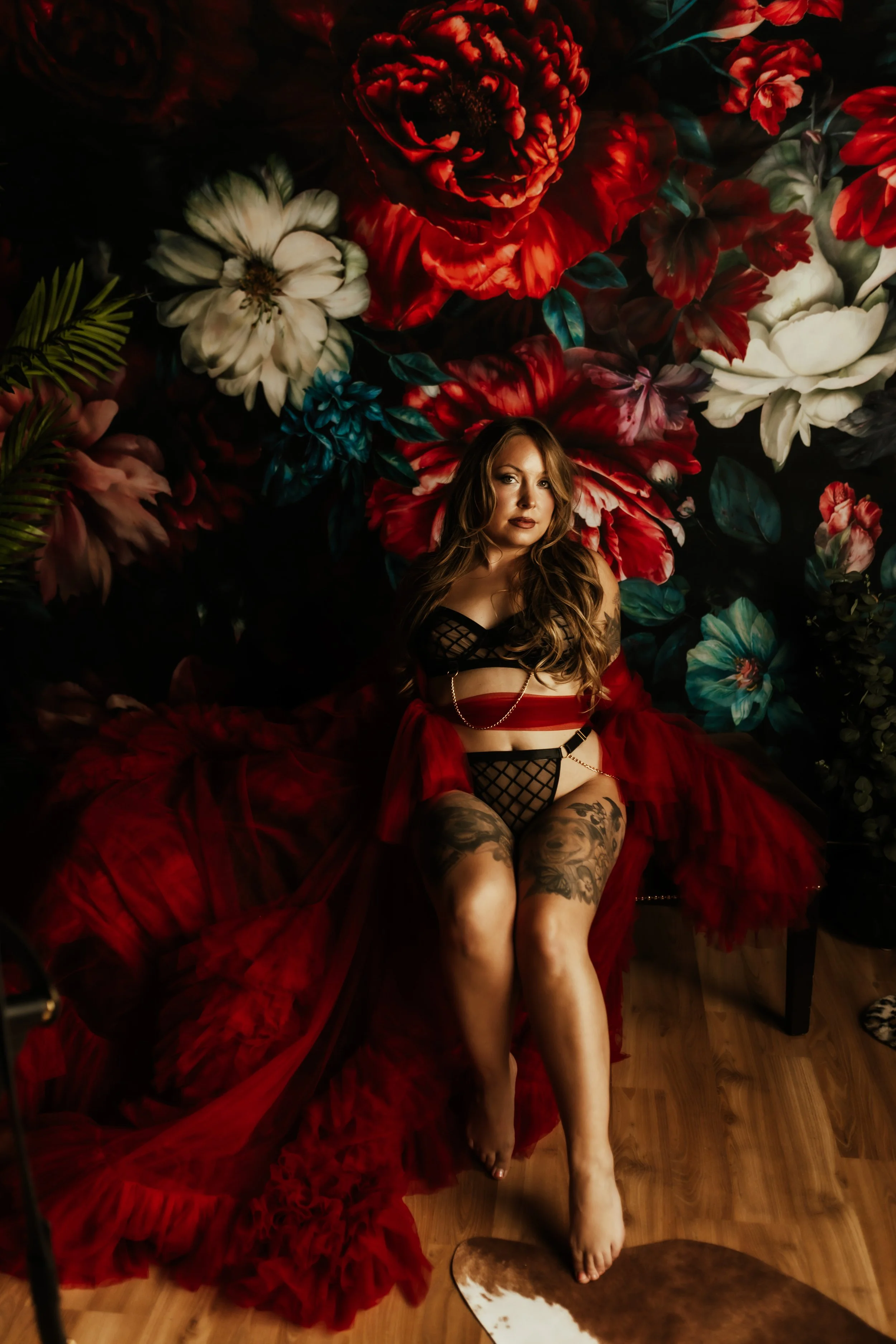 Woman with long wavy hair, tattoos on legs, wearing black fishnet lingerie and a red tulle skirt, sitting in front of a dark floral backdrop featuring large red, white, and blue flowers, with a cowhide rug on the wooden floor.