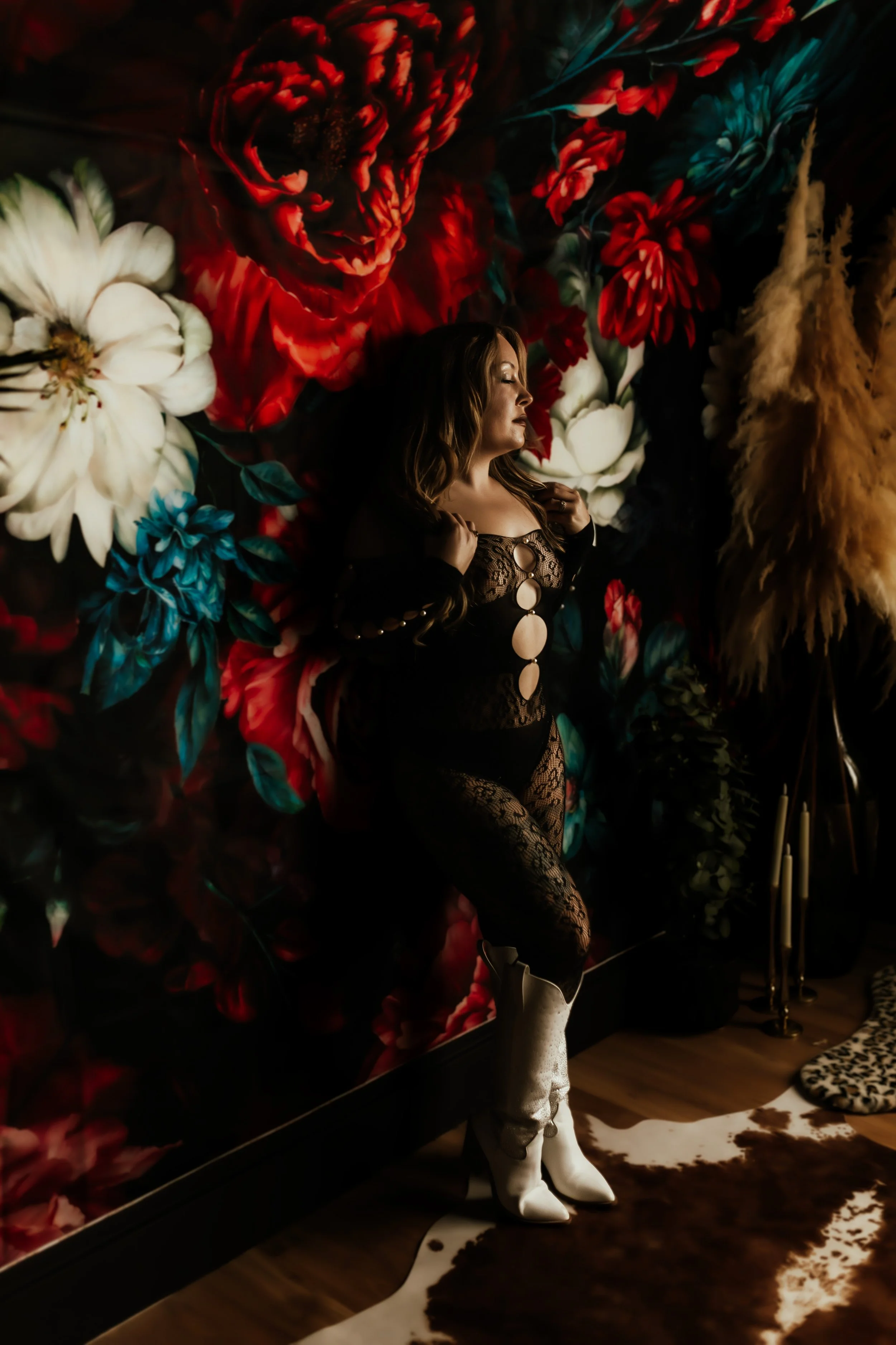 A woman with long wavy hair dressed in black lace clothing and white cowboy boots standing in front of a large floral wall with red, white, and blue flowers, and a cowhide rug on the floor.
