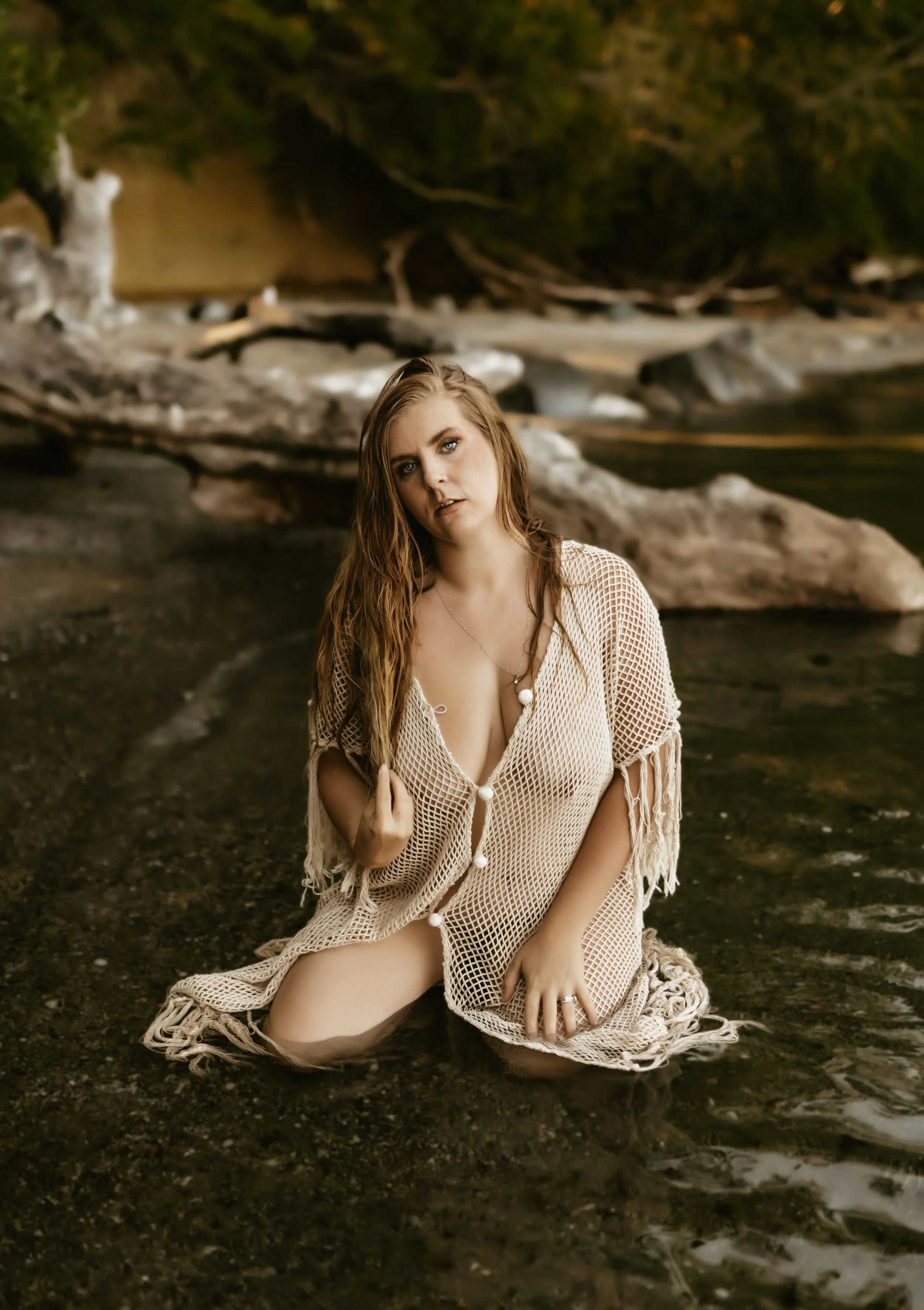 A woman with long, wavy hair kneeling in shallow water at a river, wearing a beige crocheted cover-up with a front tie, with a backdrop of rocks, driftwood, and green trees.