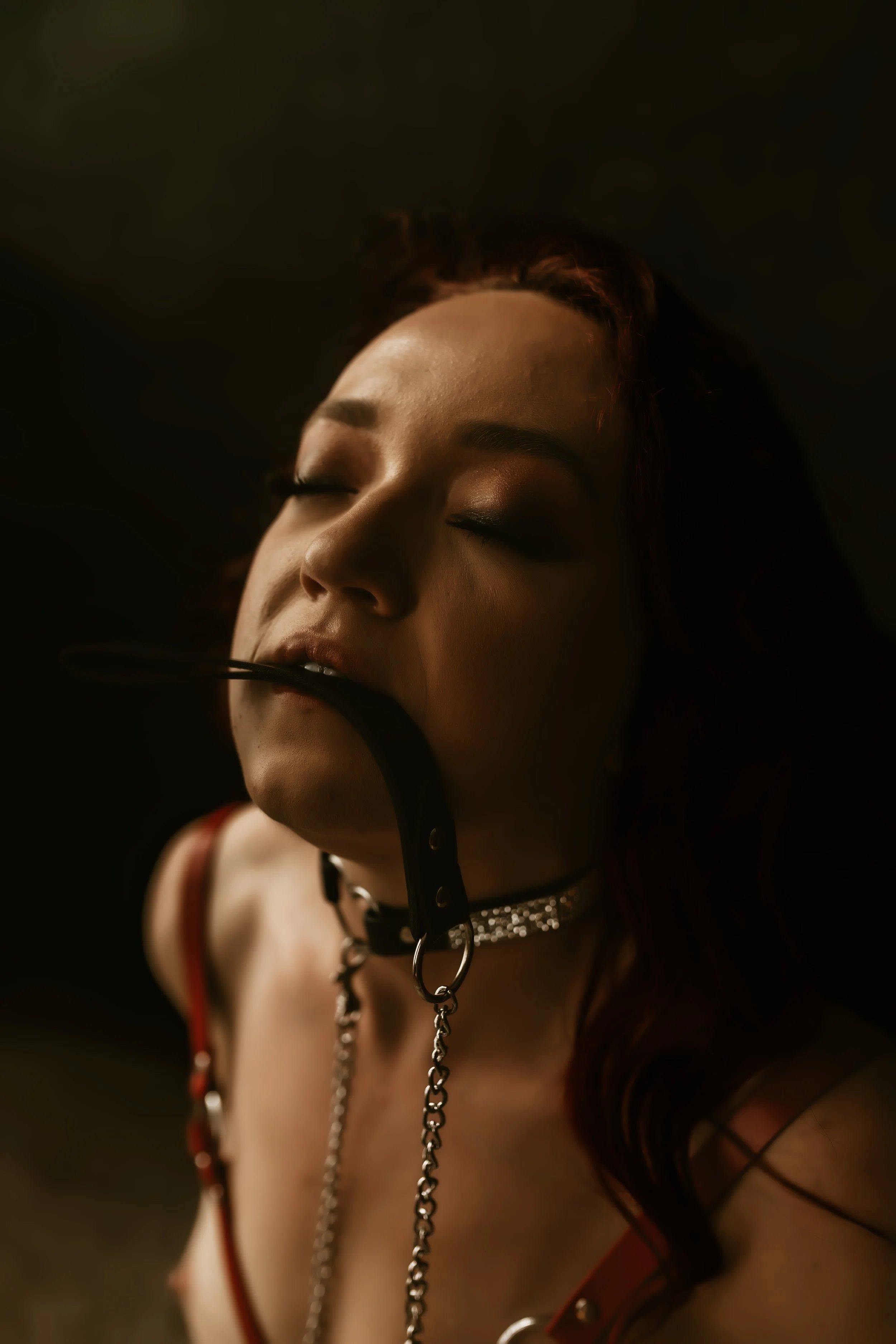 A woman with closed eyes and dark makeup wearing a collar with chains, a red and black strappy outfit, and a chain necklace, against a dark background.