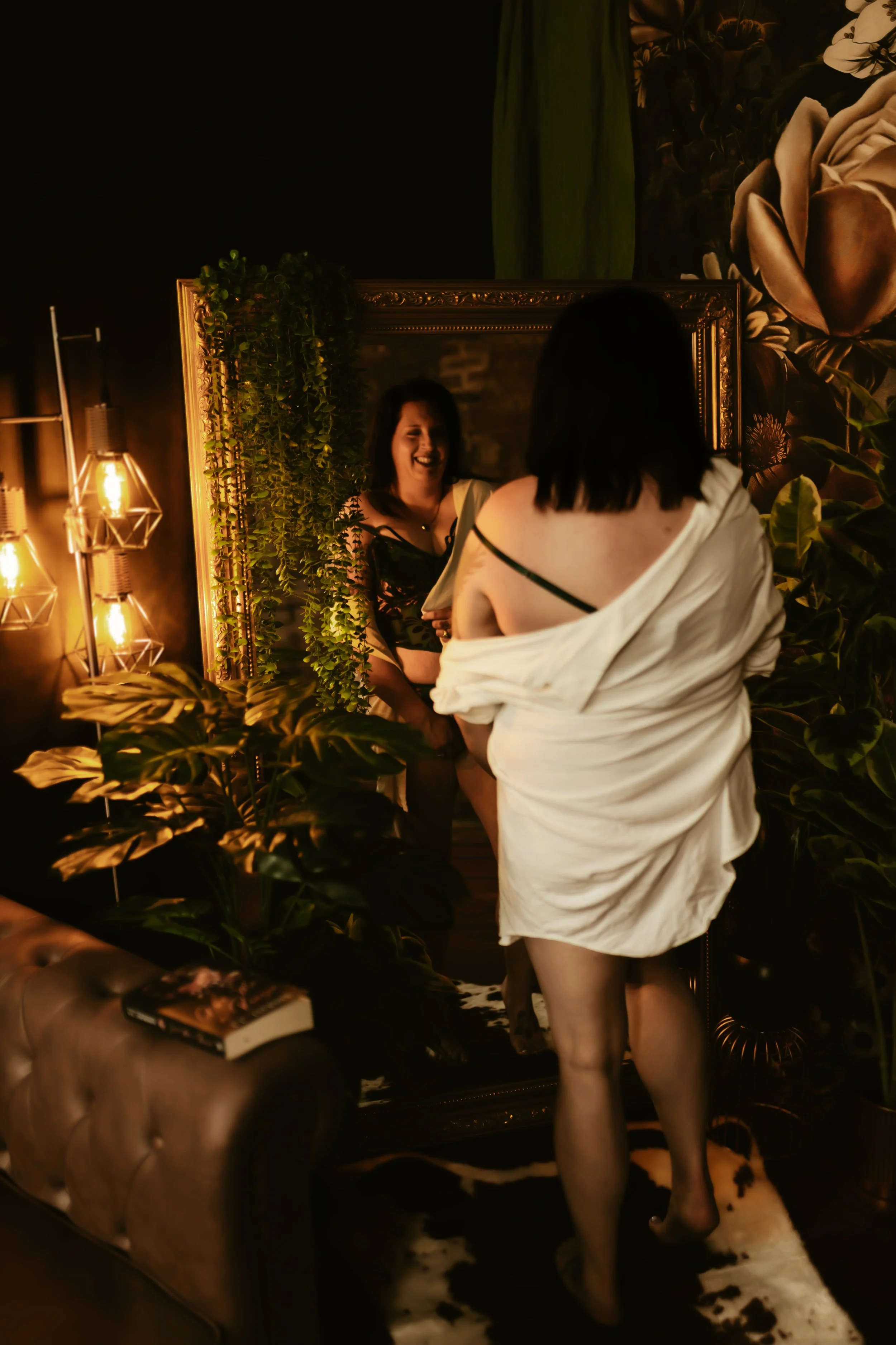 A woman stands in front of a mirror, smiling, while another woman with black hair and a white off-shoulder top, possibly a robe or dress, faces her. The room has warm lighting, green plants, and floral wall art, with a cowhide rug and a leather armch