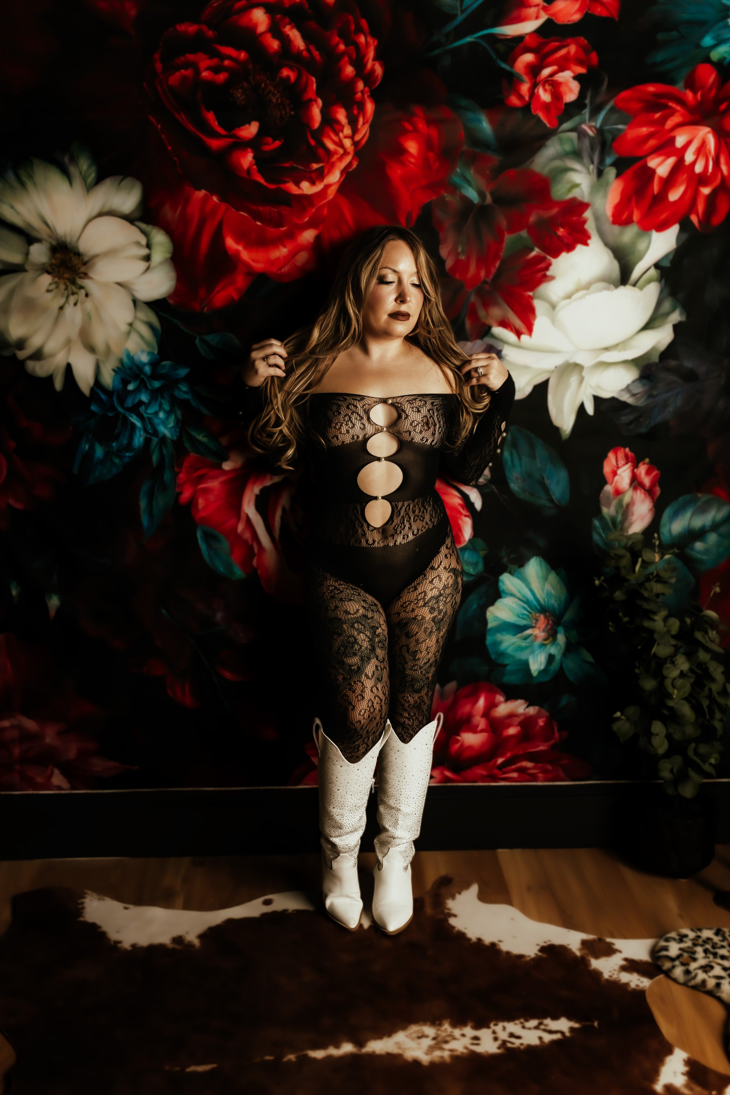 A woman in a black lace bodysuit with large circular cutouts, white cowboy boots, and long wavy hair, standing in front of a floral mural with large red, white, and teal flowers.