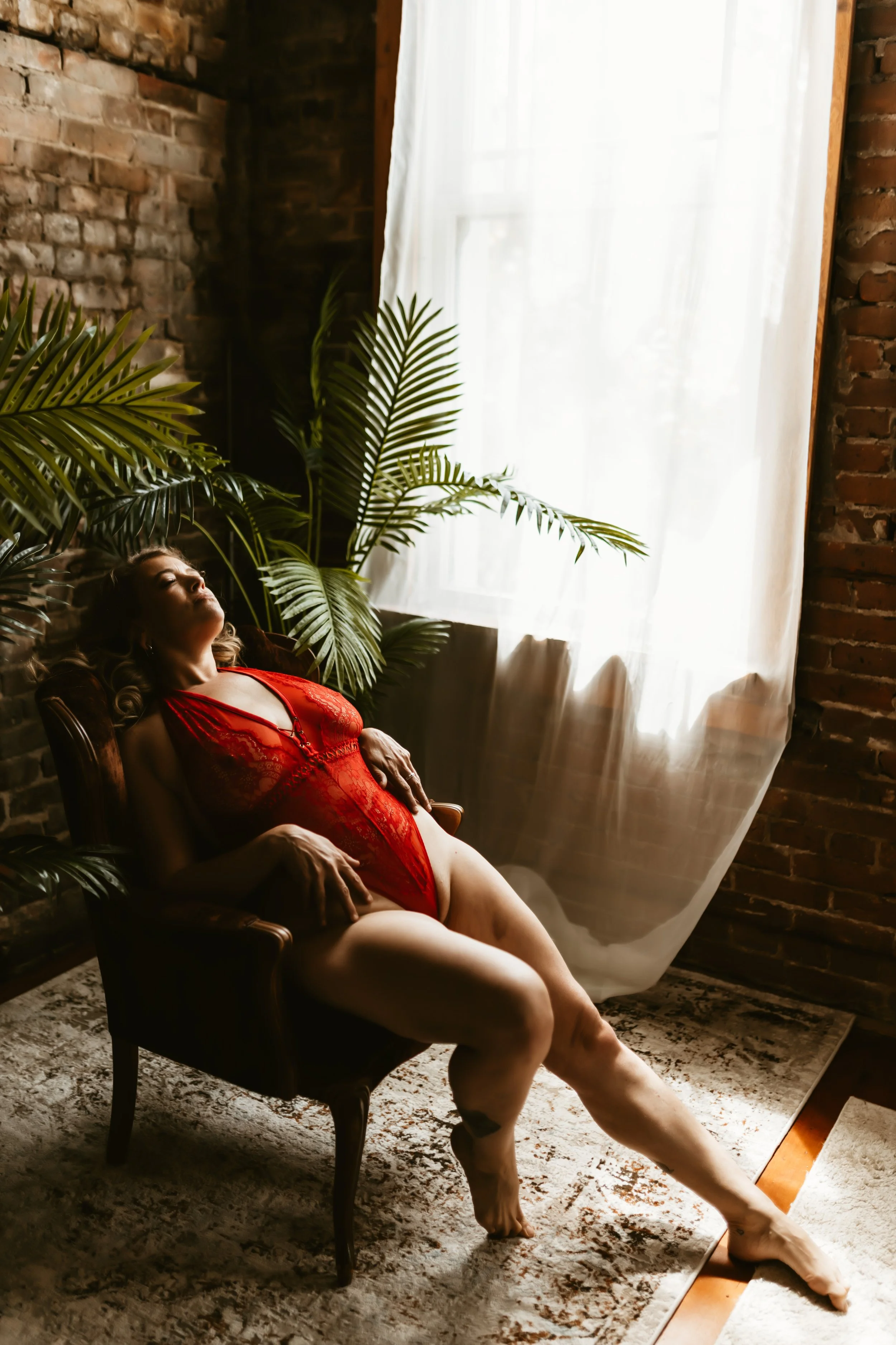 A woman in red lingerie reclining on a vintage armchair near a large window with white curtains, in a room with brick walls and tropical plants.