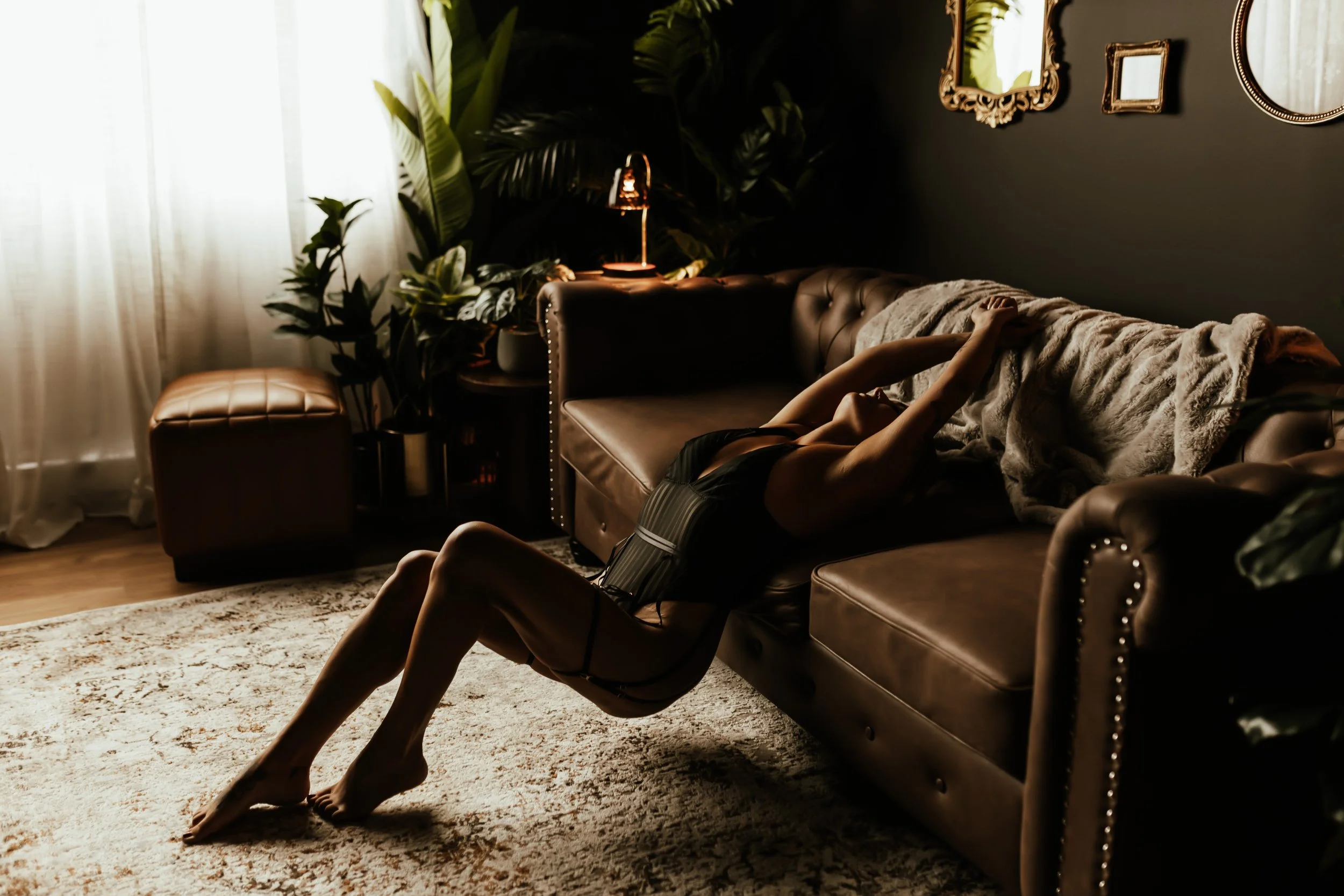 A woman lying on a leather sofa covered with a blanket in a dimly lit living room with green plants, framed mirrors on the dark wall, and soft natural light coming through sheer curtains.