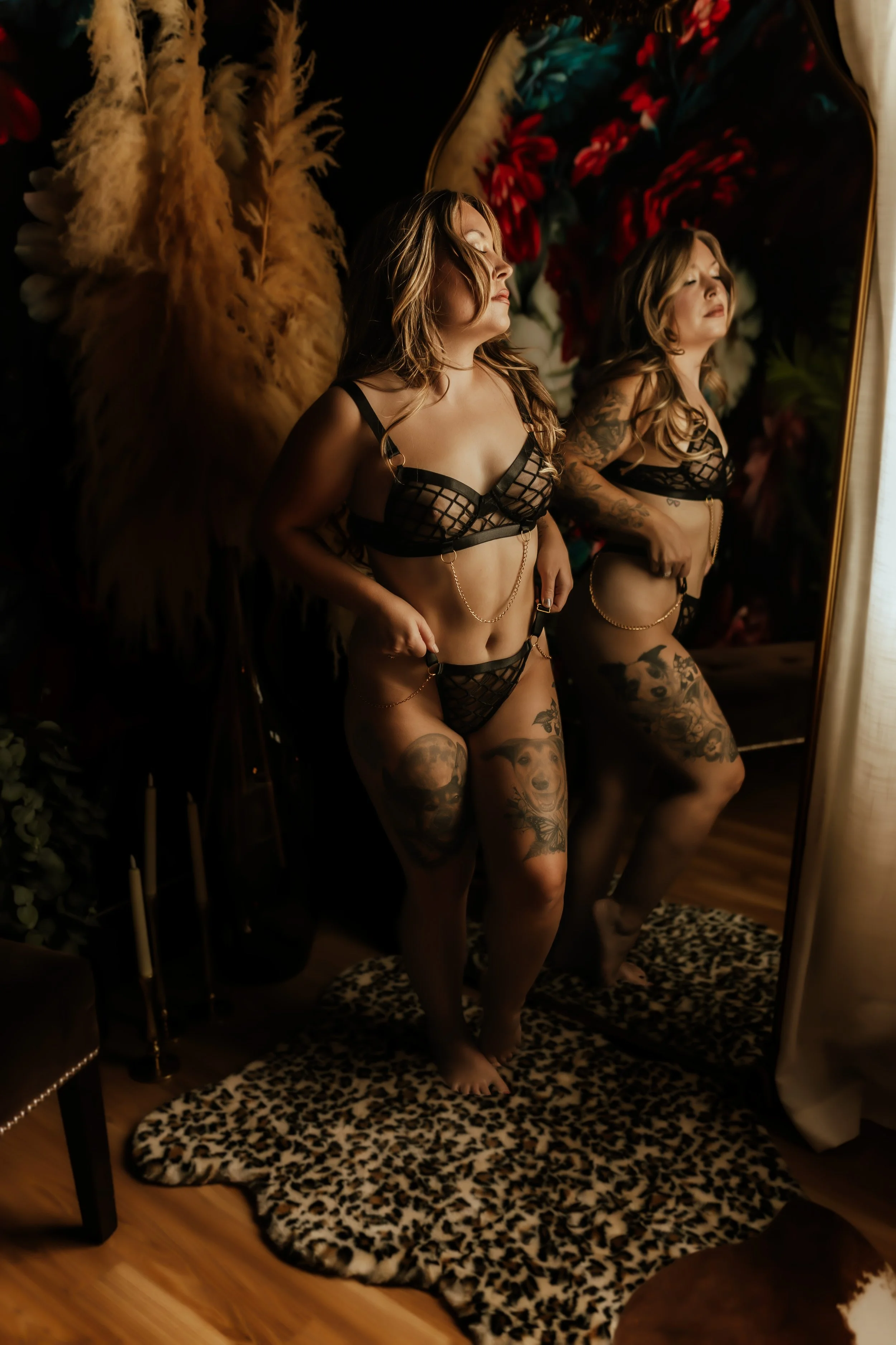 Woman in black lingerie standing in front of a mirror, with tattoos on her legs and arms, in a room with dark floral wallpaper, a leopard print rug, and pampas grass decor.