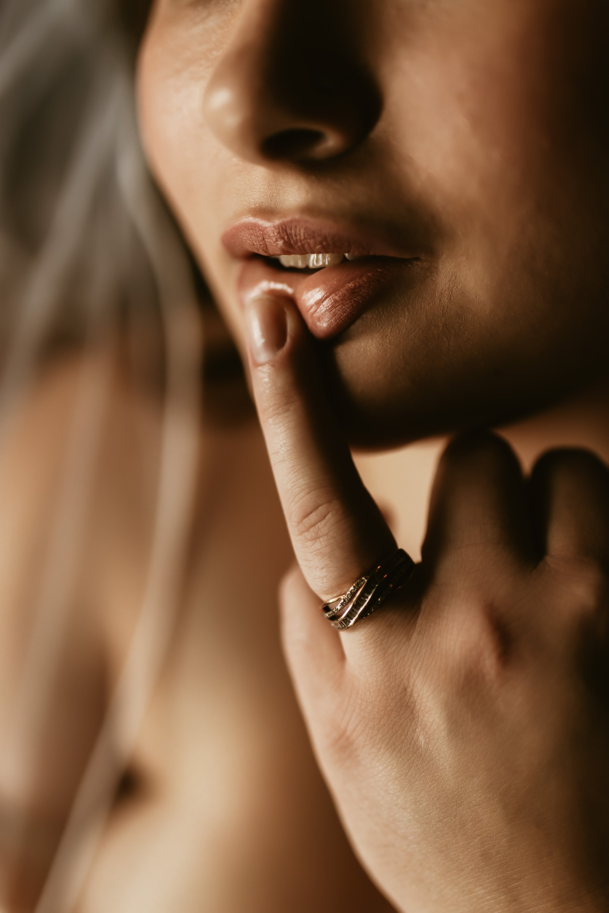 Close-up of a woman's face, showing her lips, chin, and part of her nose, with her finger touching her lips, wearing rings on her finger.