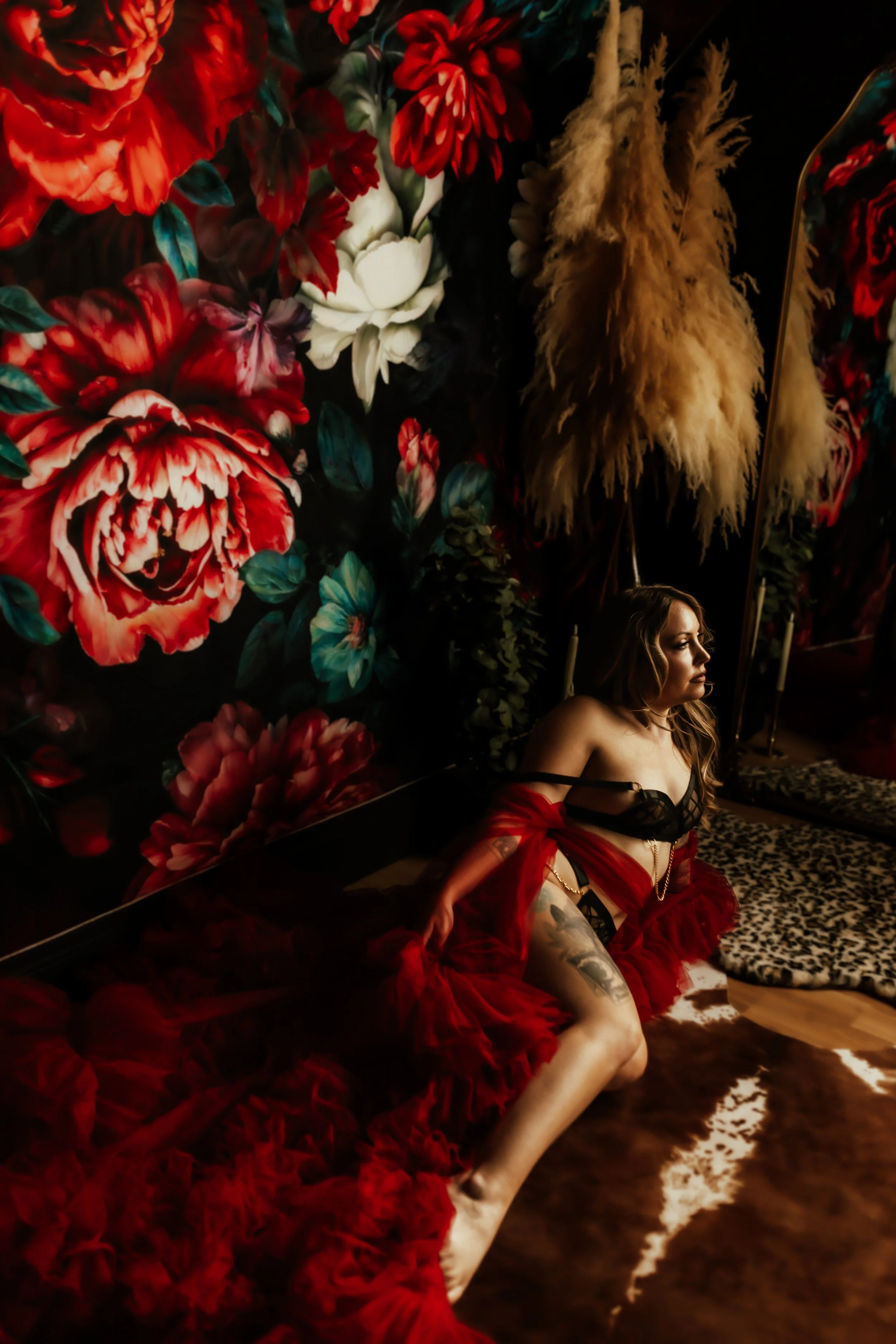 A woman with long hair and tattoos sitting on the floor, wearing black lingerie and a red sheer robe, in front of a large floral wall with red, white, and dark flowers and dried pampas grass.