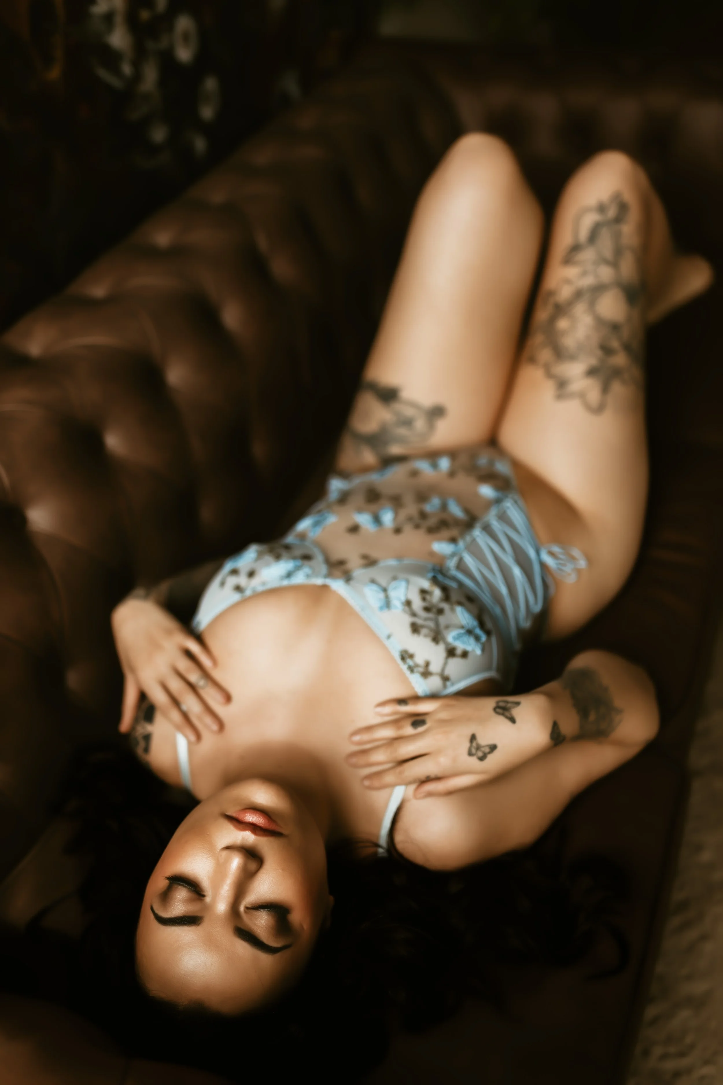 A woman with dark hair and tattoos on her arms is lying on a dark leather couch with her eyes closed. She is wearing a light-colored lace top and patterned shorts. Her left hand is on her chest, and her right hand is on her head.