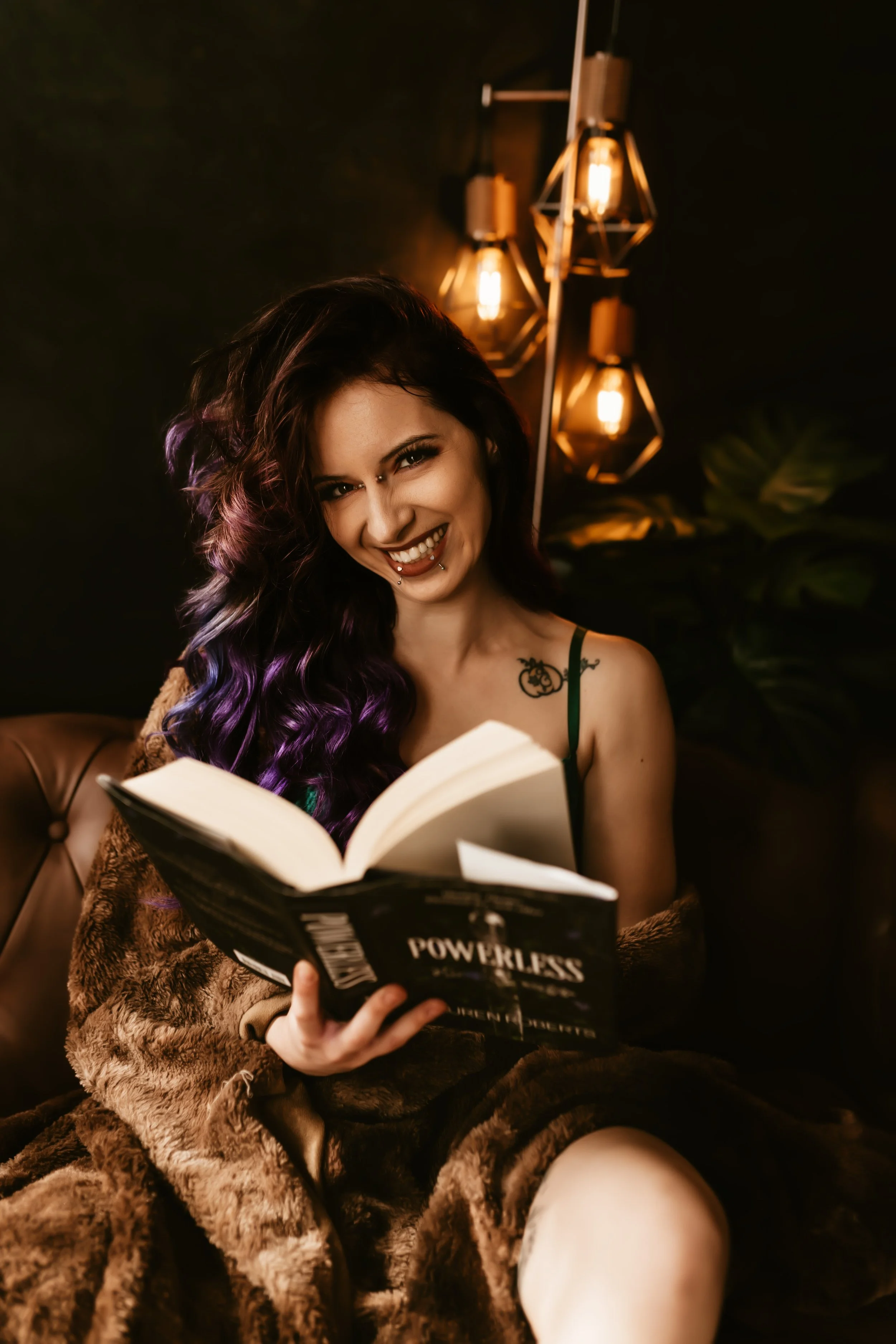 A smiling woman with dyed purple hair and piercings reading a book titled 'Powerless' in a cozy, dimly lit room with warm lighting and a dark background.