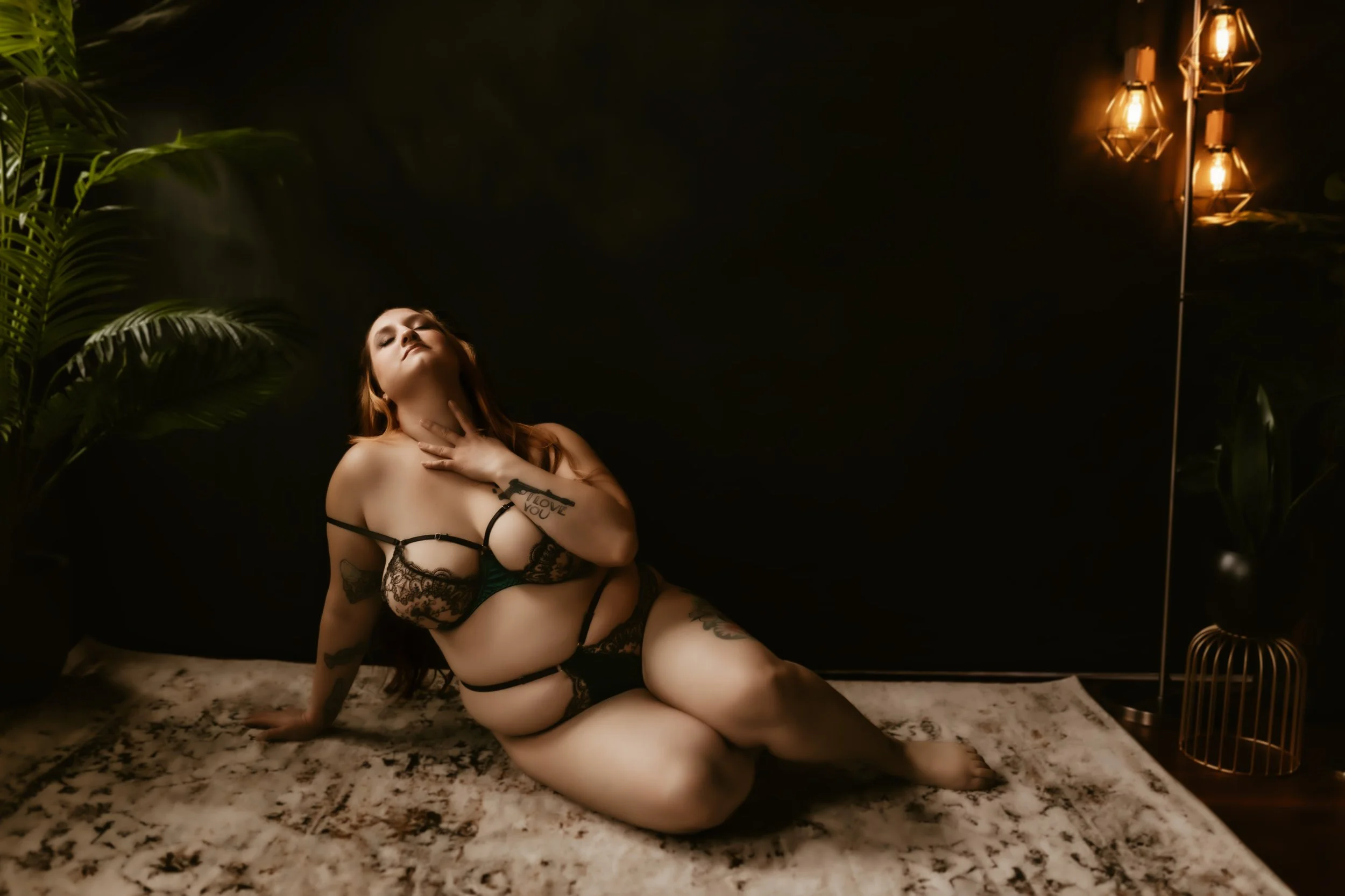 A woman with tattoos posing on a carpeted floor in lingerie, surrounded by green plants and lit by warm ceiling lights, with a dark background.