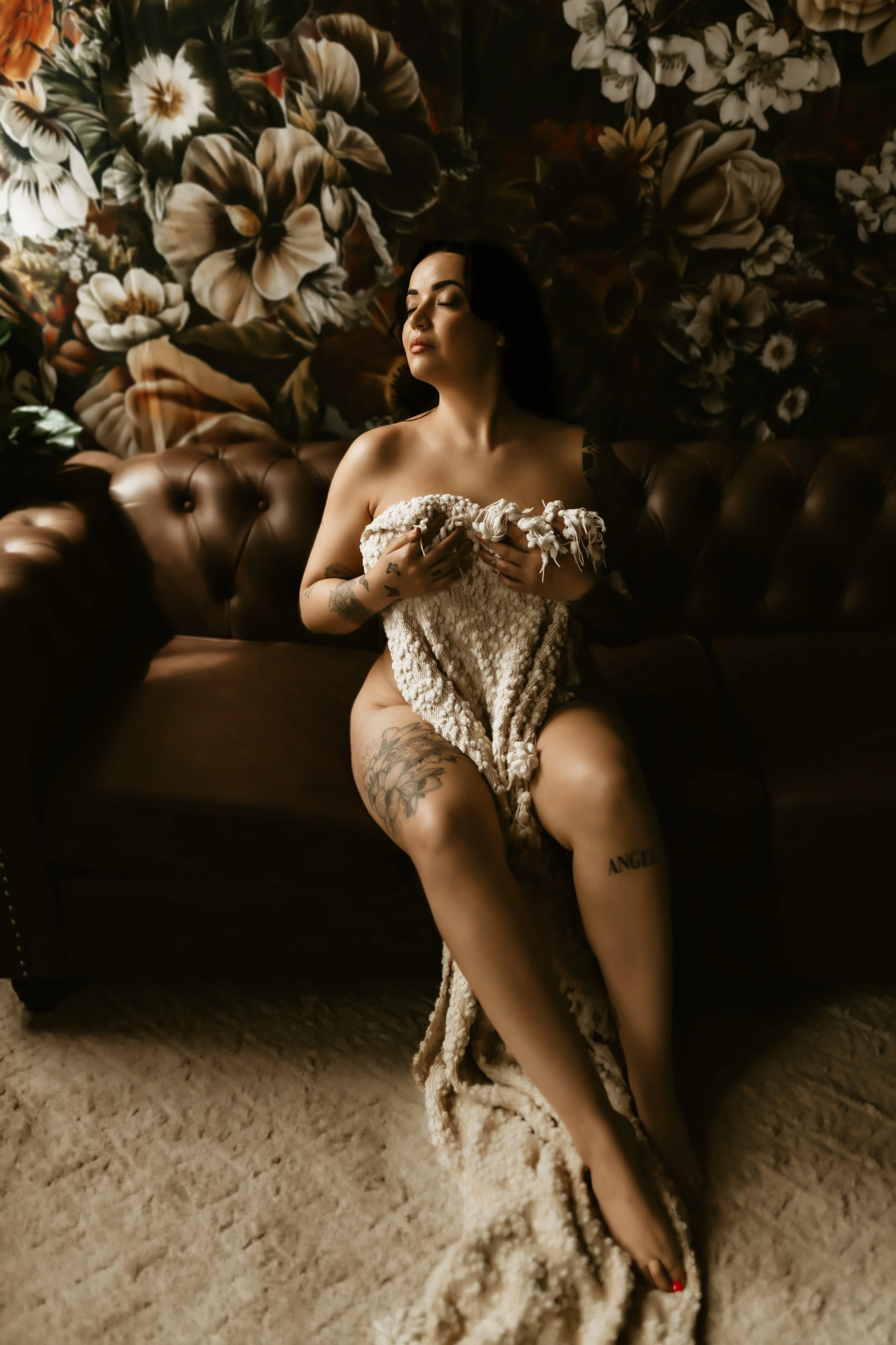 A woman with dark hair and tattoos on her arm and leg is sitting on a beige carpeted floor, holding a fluffy beige blanket or towel against her chest with her eyes closed. She is nude from the waist up and appears to be in a relaxed or contemplative 