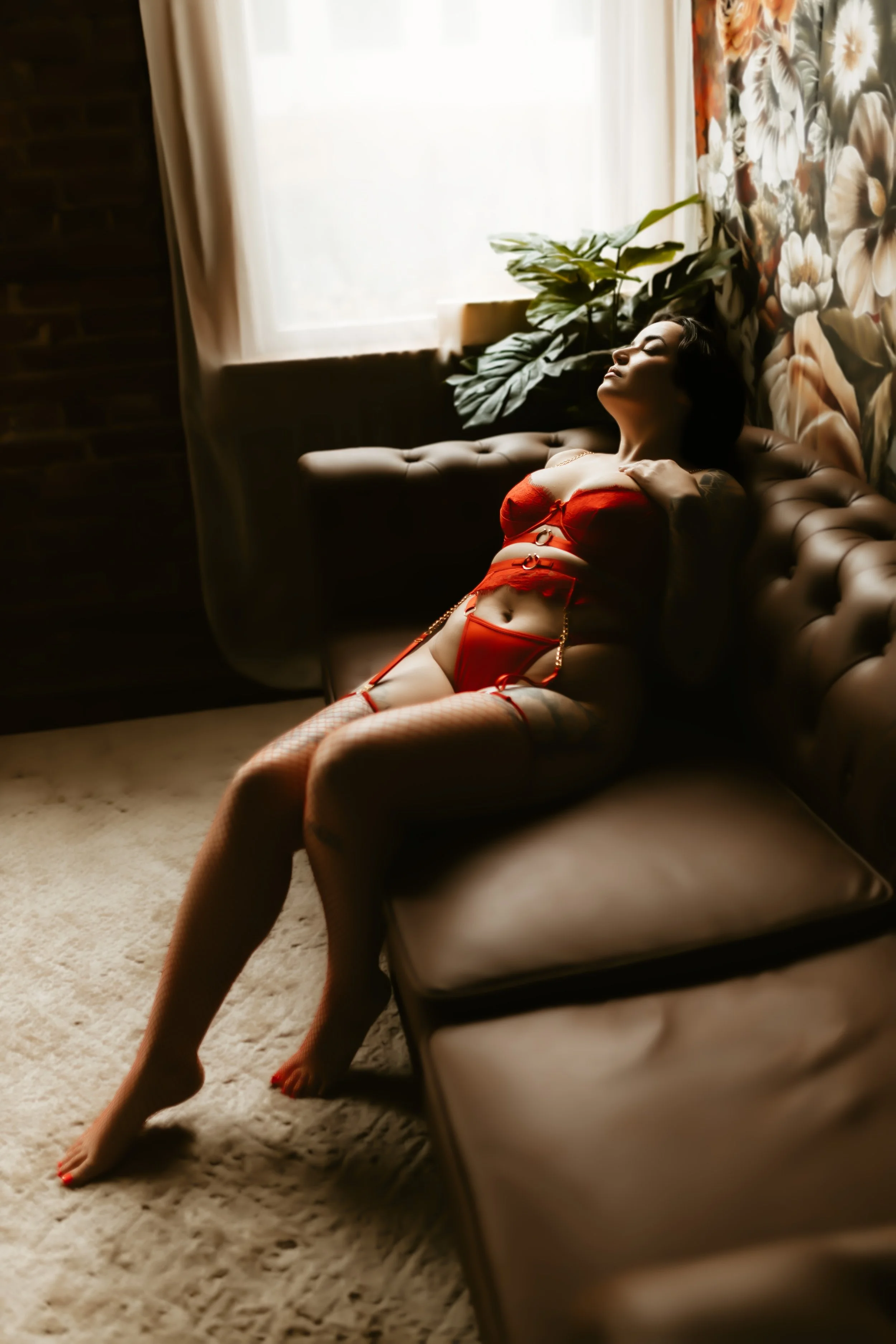 Woman in red lingerie and fishnet stockings lying on a beige couch in a dimly lit room with floral wallpaper, a large window with curtains, and a potted plant nearby.