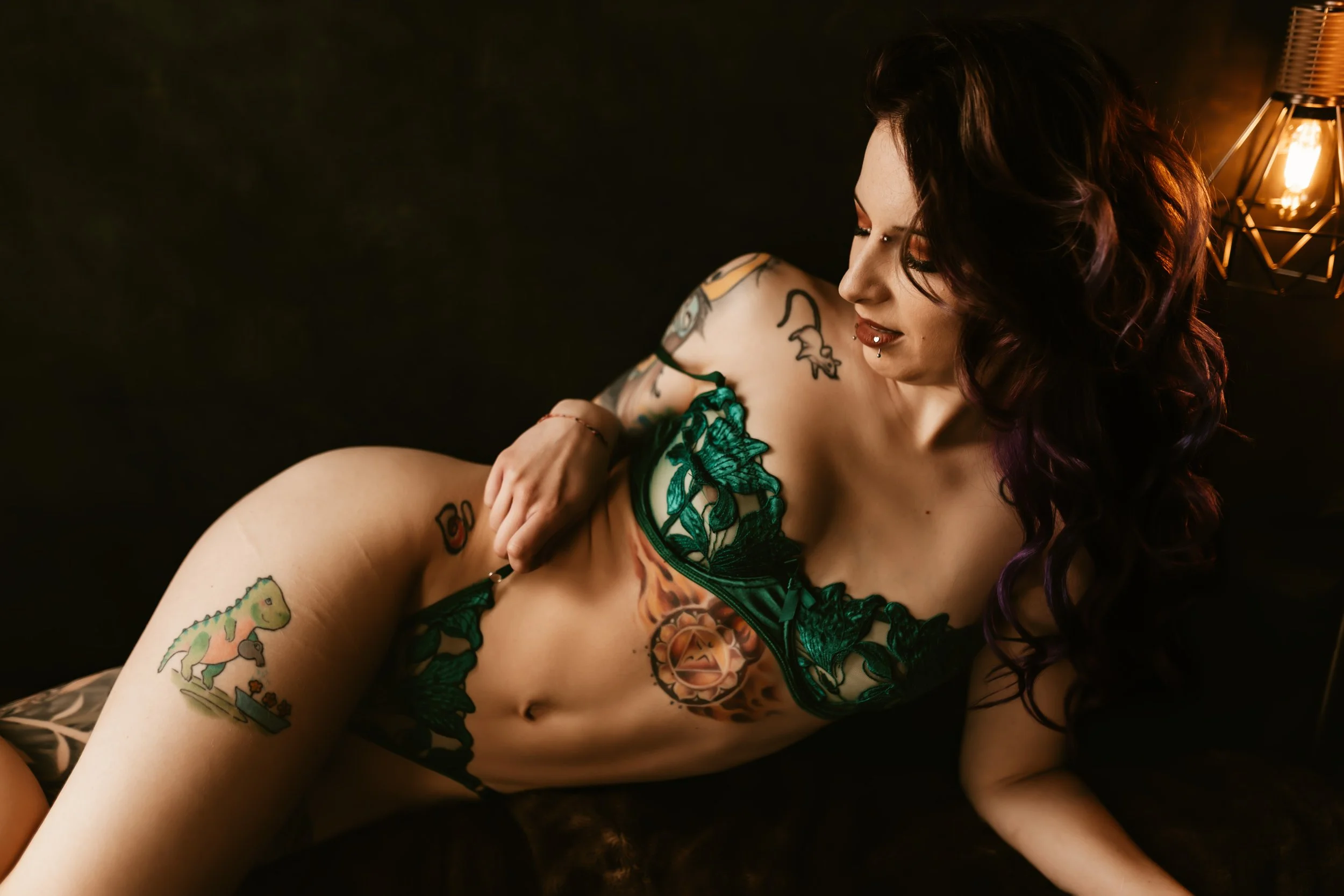 A woman with colorful tattoos on her arm and torso, dark wavy hair, and green lingerie, lying on her side near a lamp with a warm orange light.