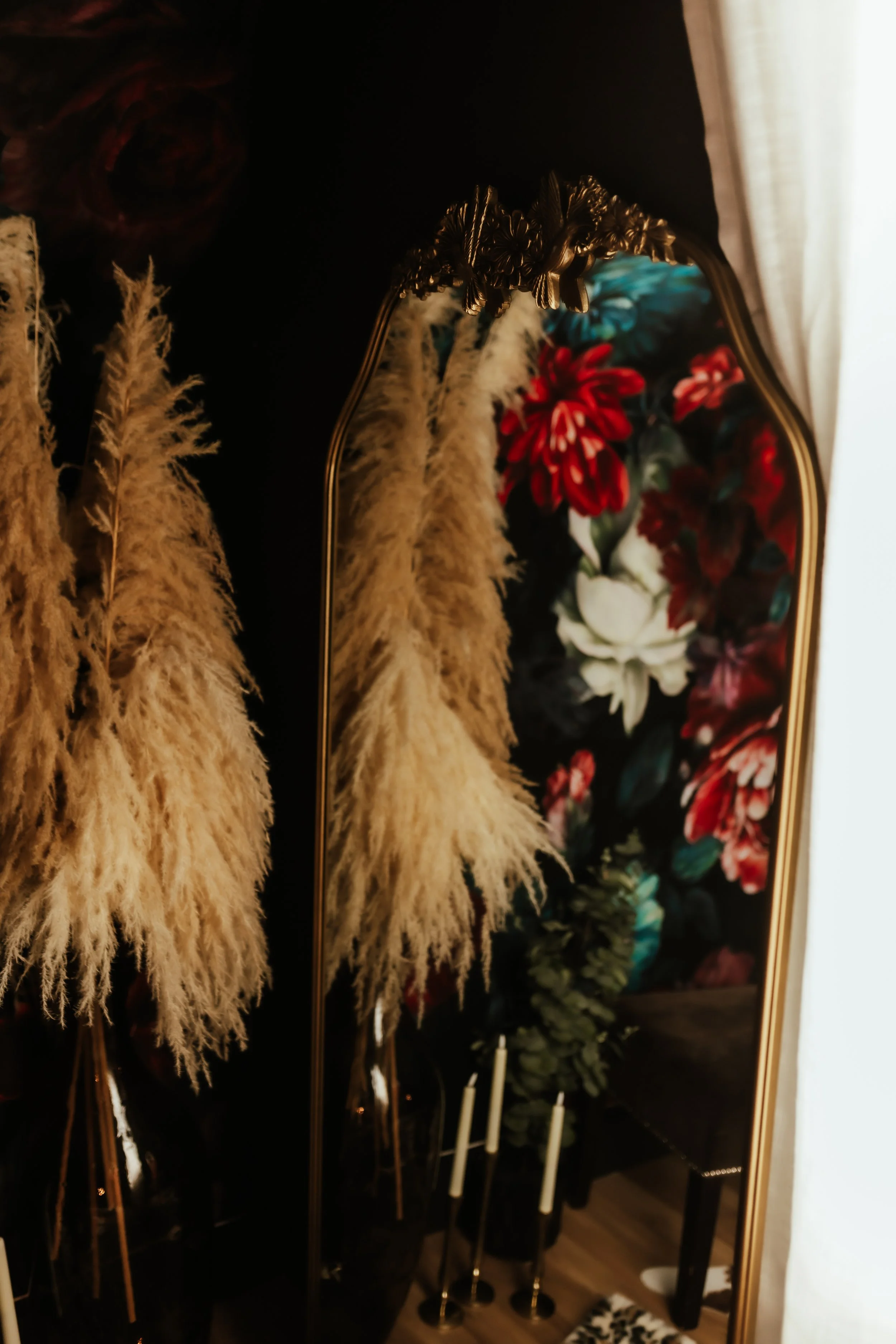 A decorative mirror with a floral design, showing inside a room with dried pampas grass and red and white flowers.