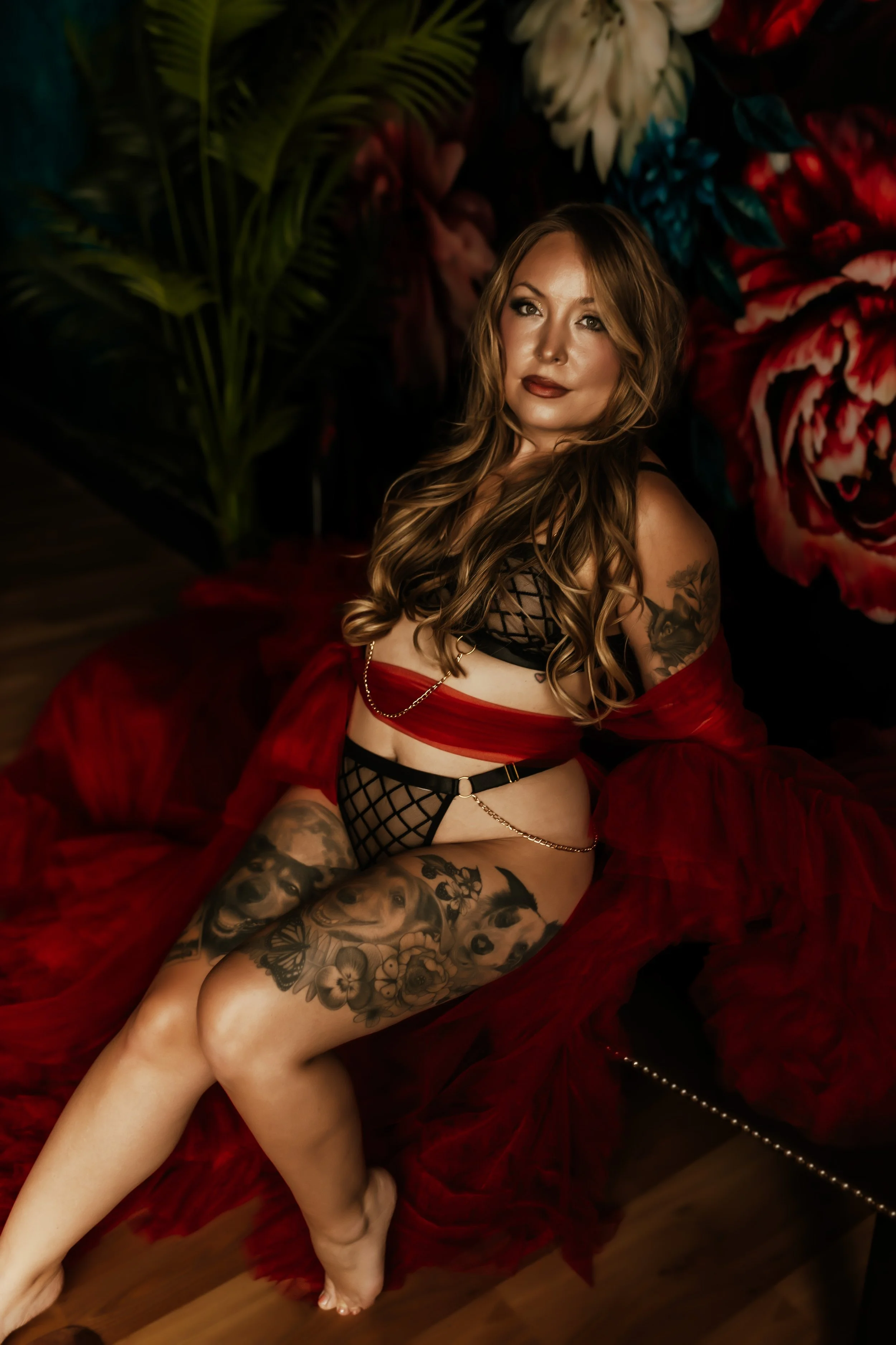 A woman with long, wavy hair and tattoos sitting on a red velvet surface, posing in black fishnet lingerie and a red sheer robe, with a background of dark floral and tropical leaves.