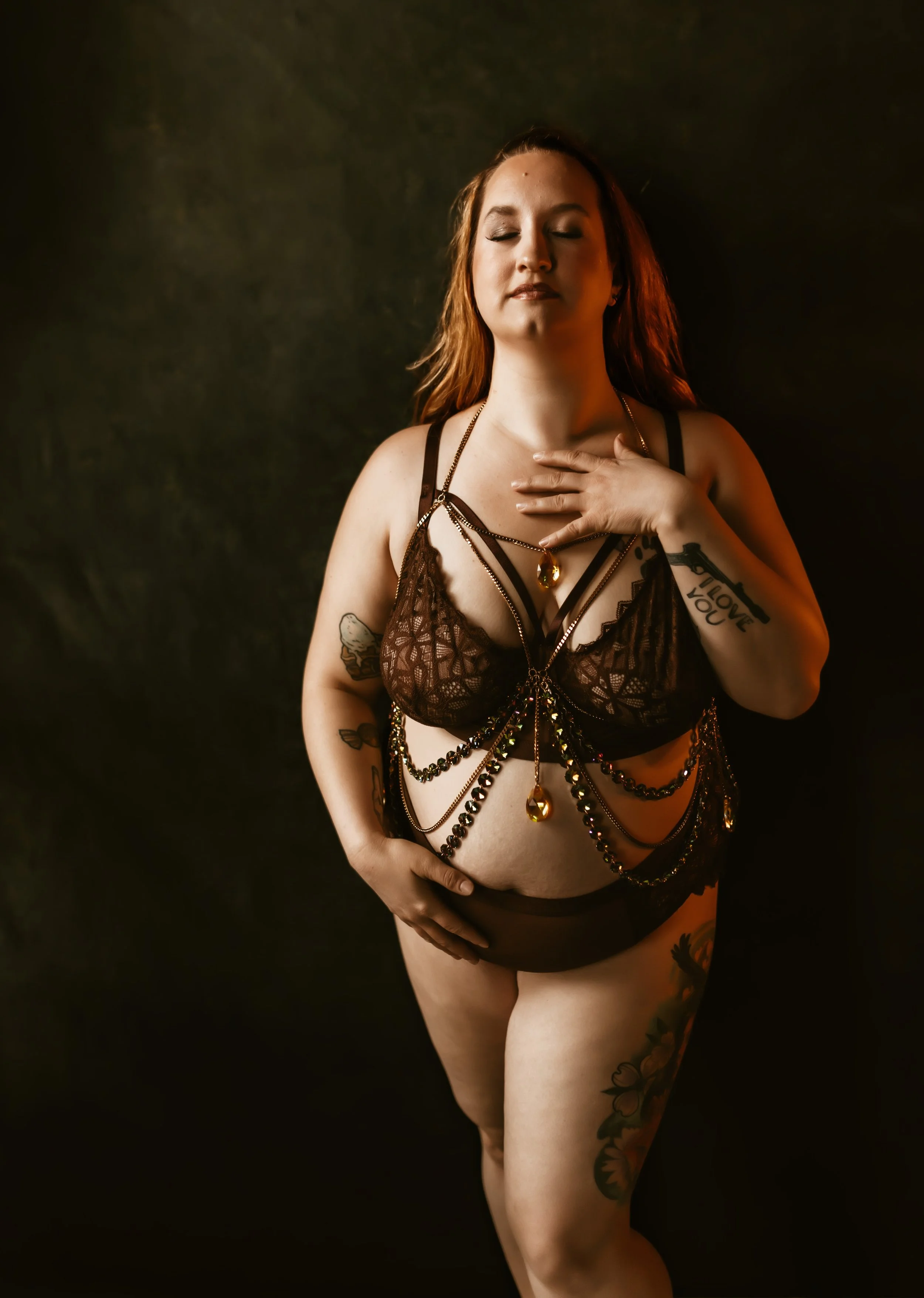Pregnant woman in black lace lingerie with layered necklaces and tattoos on her arm and thigh, standing against a black background with eyes closed and hand on chest.