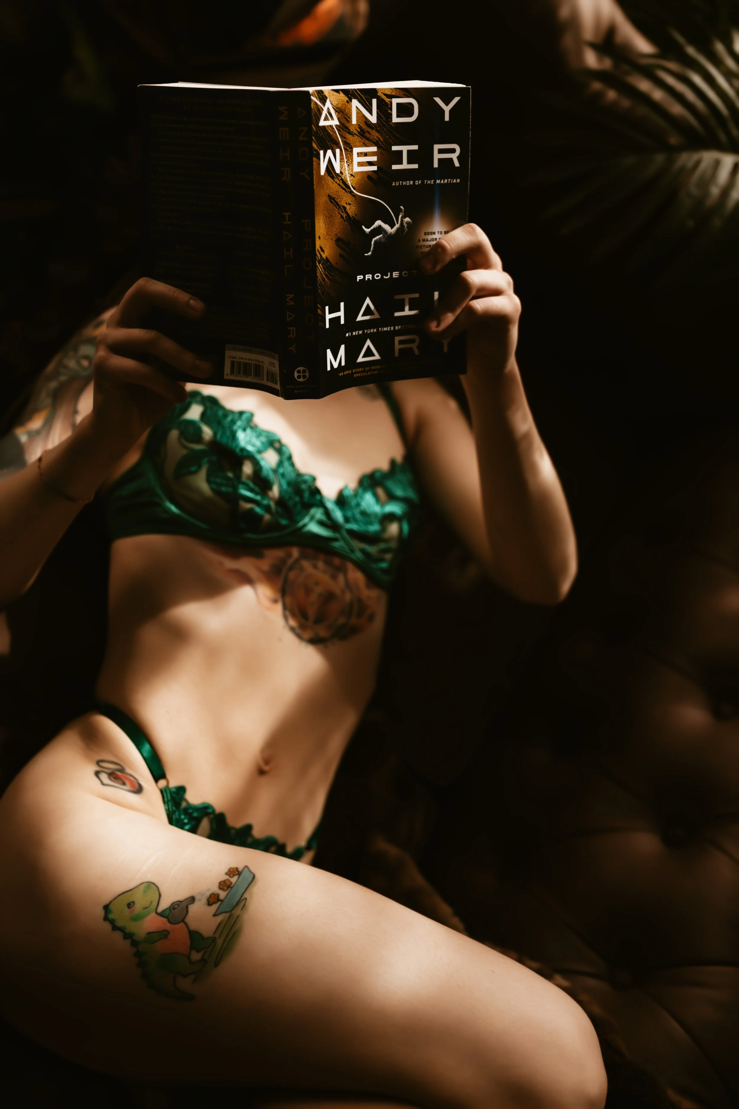 Person with tattoos wearing green lingerie reading a book titled 'The Martian' in a dimly lit environment.