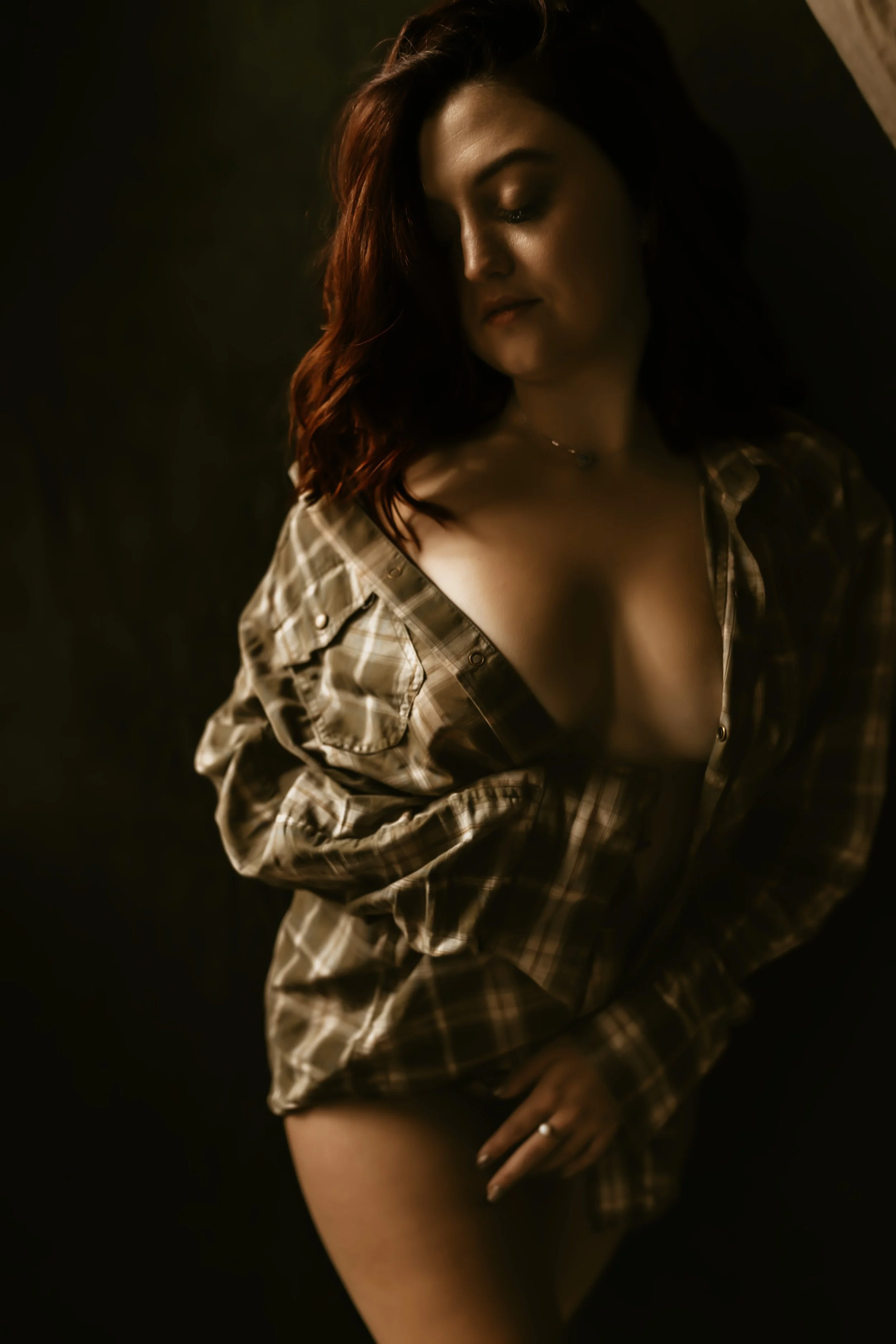 A woman with long, wavy brown hair, wearing a checkered shirt partly unbuttoned, showing her shoulder and thigh, against a dark background.