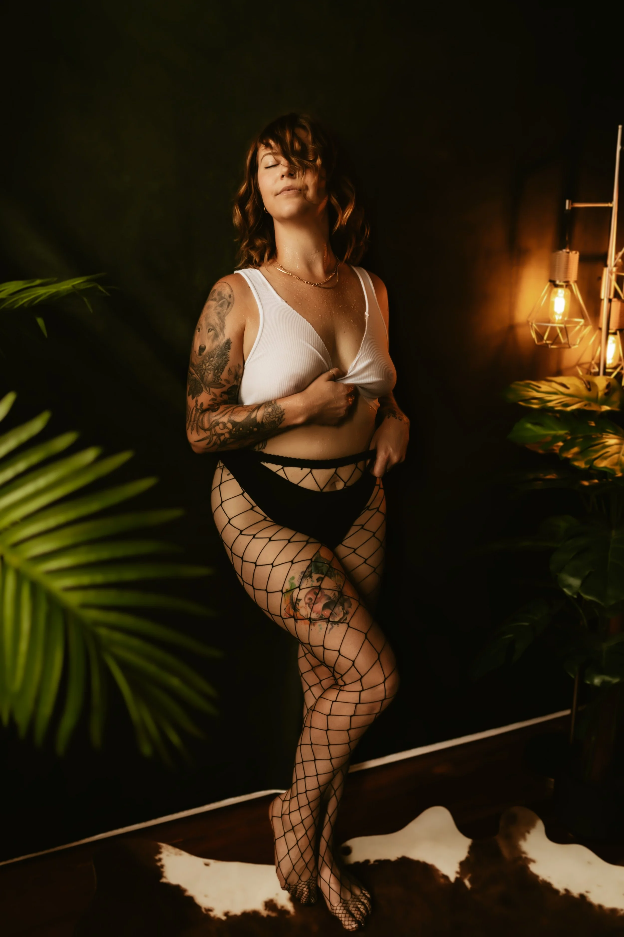 A woman with shoulder-length brown hair, tattoos on her left arm and leg, stands against a dark background with warm lighting. She is wearing a white tank top, fishnet stockings, and jewelry, with a relaxed pose and eyes closed.