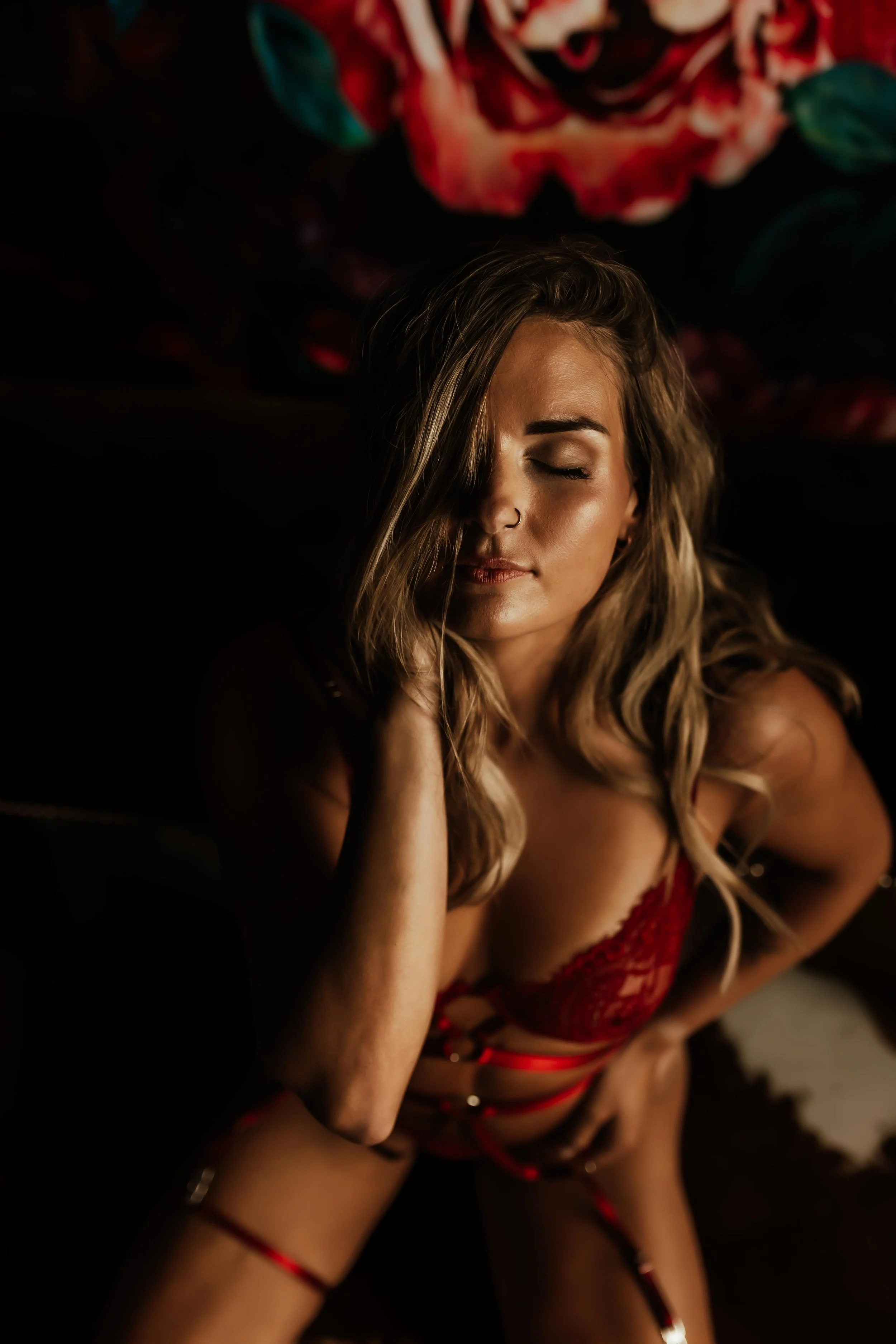 A woman with long wavy hair, wearing red lingerie with straps, sitting in a dark room with her eyes closed, surrounded by a dark background with a large floral mural above her.