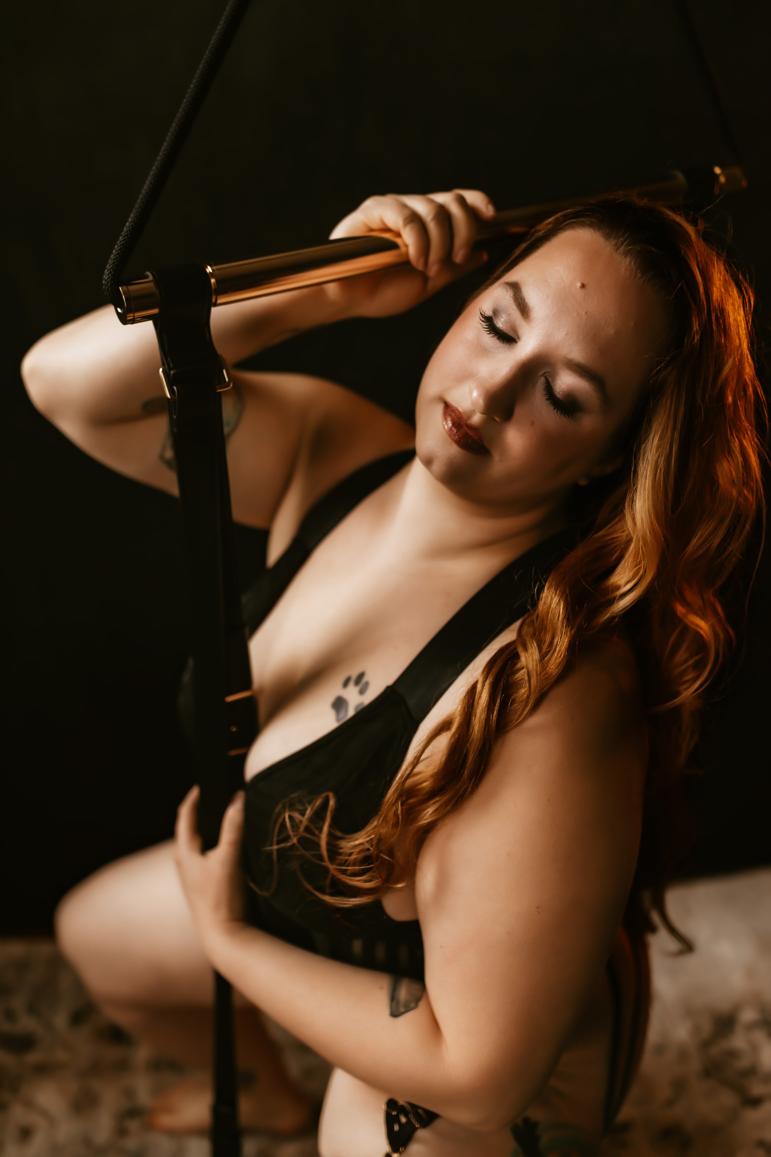 A woman with long red hair and tattoos on her arm, wearing black lingerie, holding a microphone stand, with her eyes closed, in a dimly lit room.