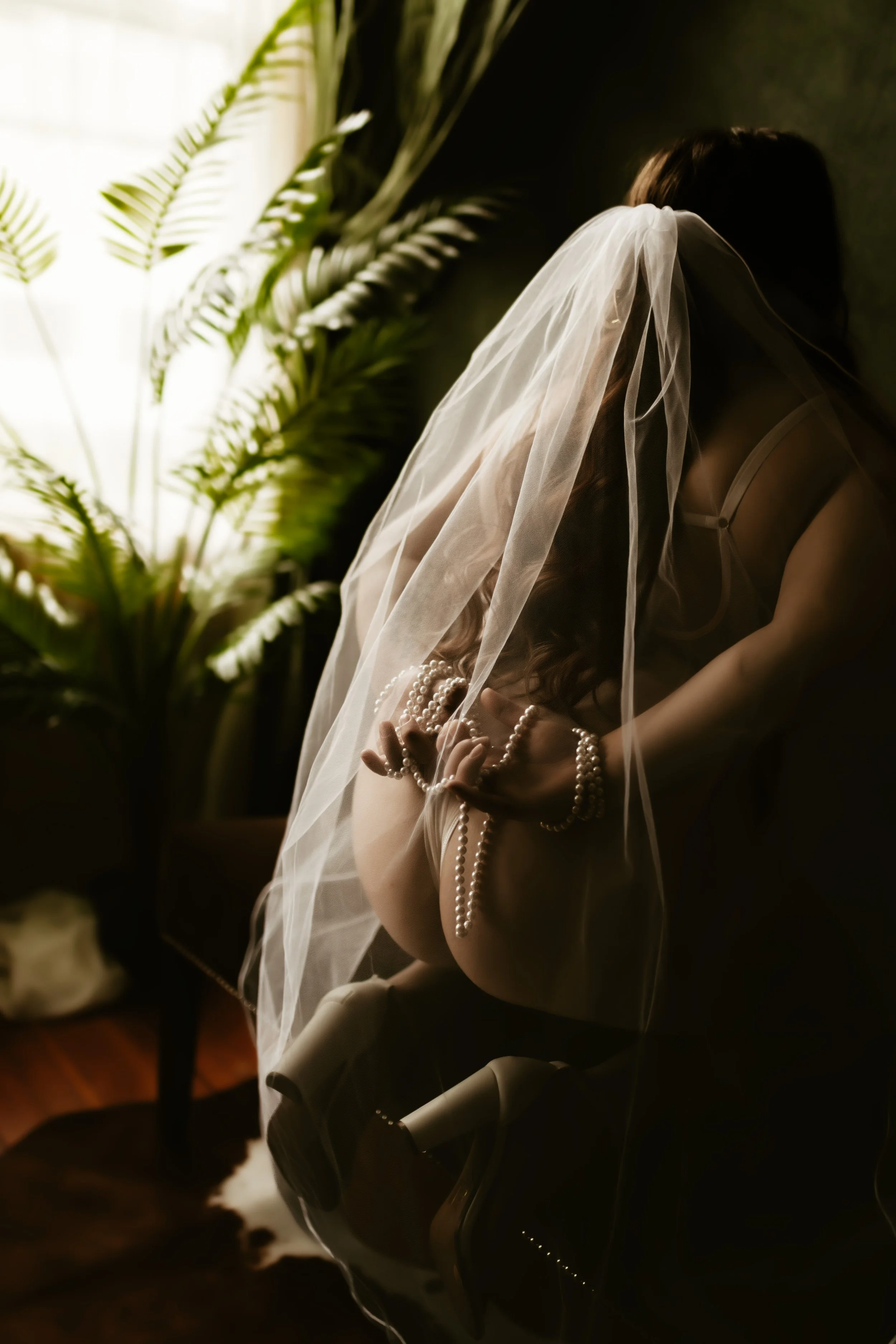 A woman wearing a wedding veil and pearl jewelry is sitting in a dark room near a window with sunlight coming through, with large green plants behind her.