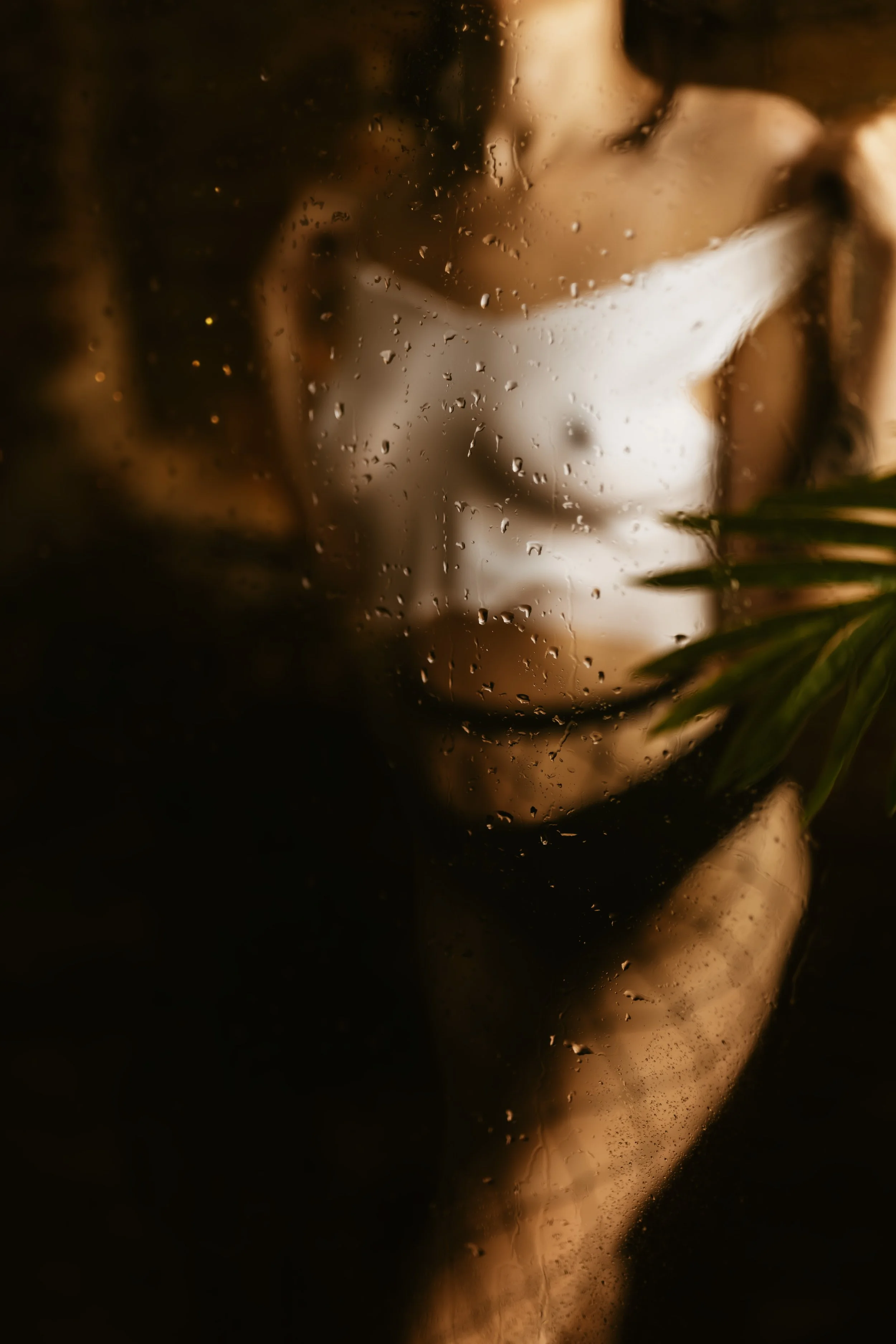 A person with long hair in a ponytail, seen through a rain-covered window, with a beverage mug nearby.