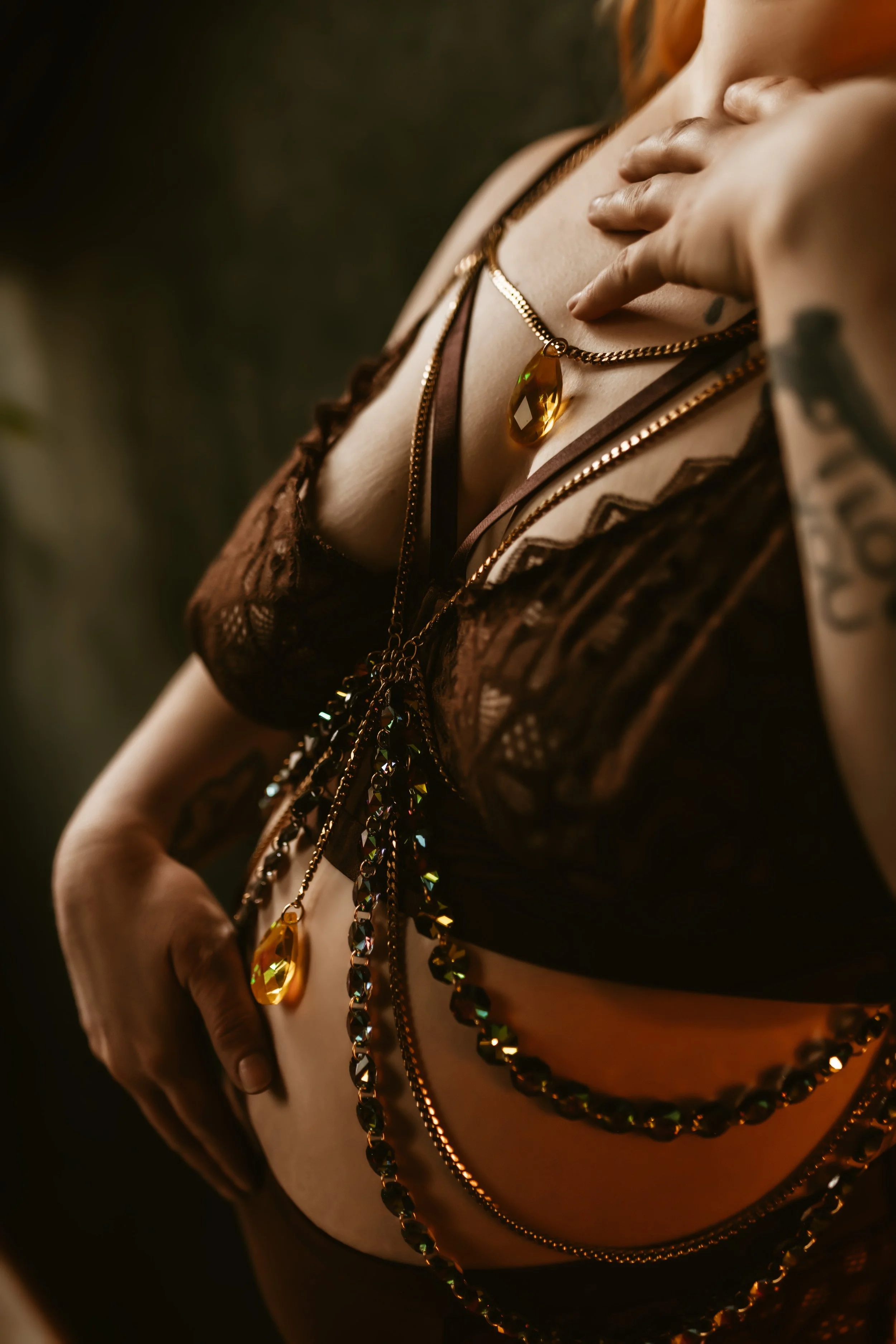 Close-up of a woman wearing layered gold and black jewelry with a large amber gemstone pendant necklace, a lace brown top, and visible tattoos on her arm, against a dark background.