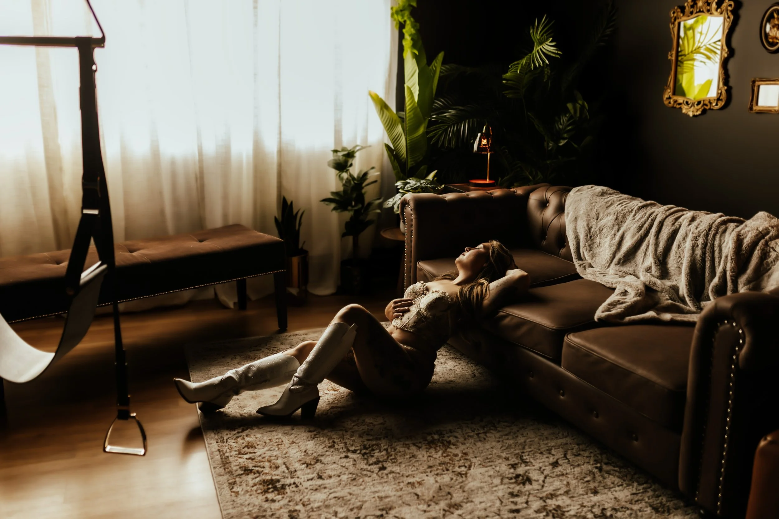 A woman relaxing on a leather couch in a dimly lit living room with large green plants, a beige blanket, and ornate mirrors on the dark wall.