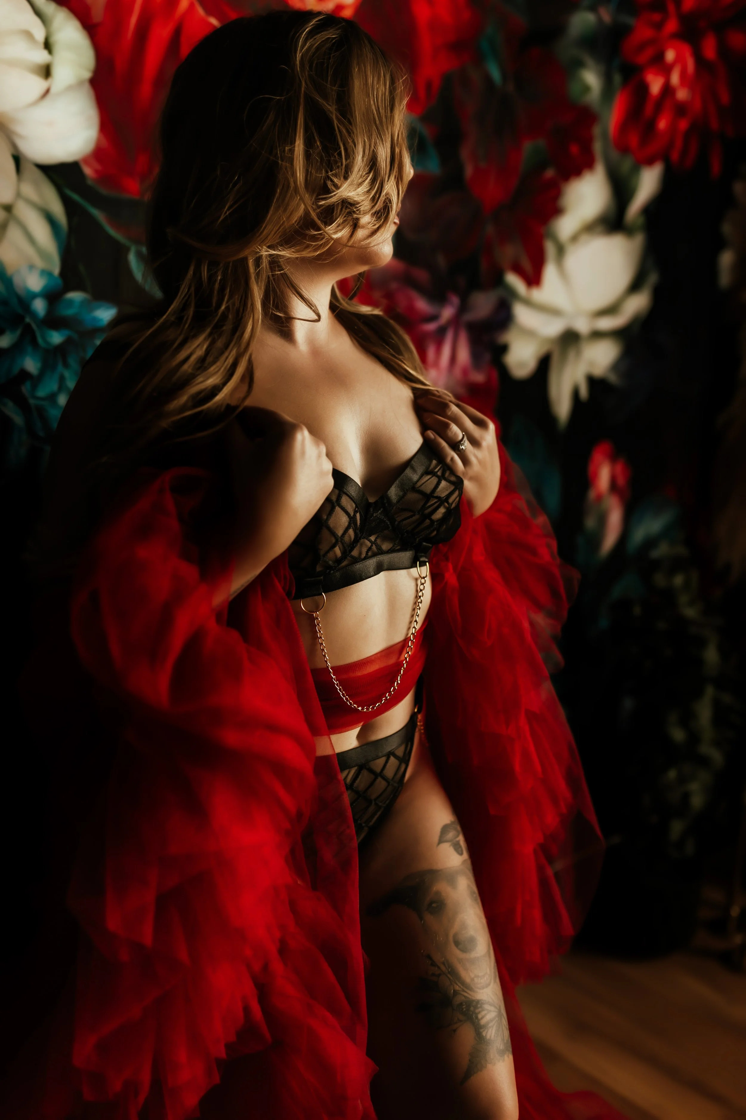 A woman with long, wavy hair and tattoos on her arm, standing in front of a wall decorated with large red, white, and teal flowers, wearing a black lingerie top, red sheer robe, and red skirt.