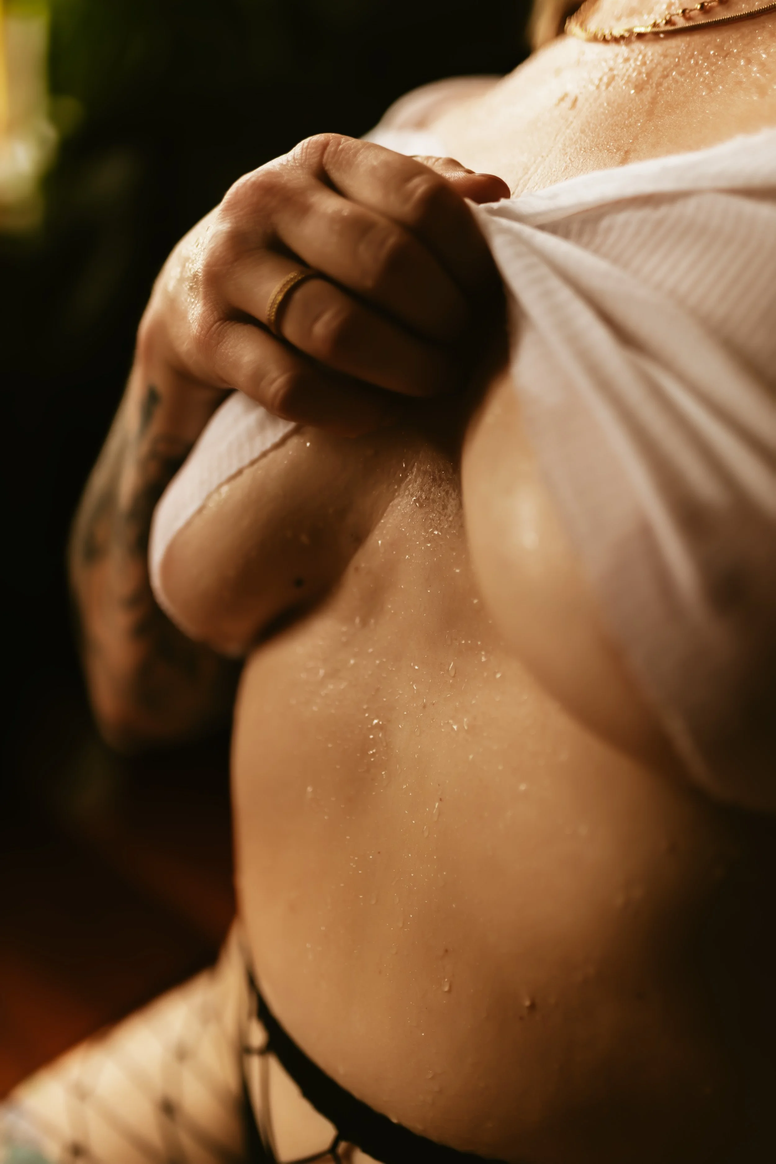 Close-up of a person's shoulder and neck, with water droplets on skin, wearing a white fabric garment and gold jewelry, in a dimly lit setting.