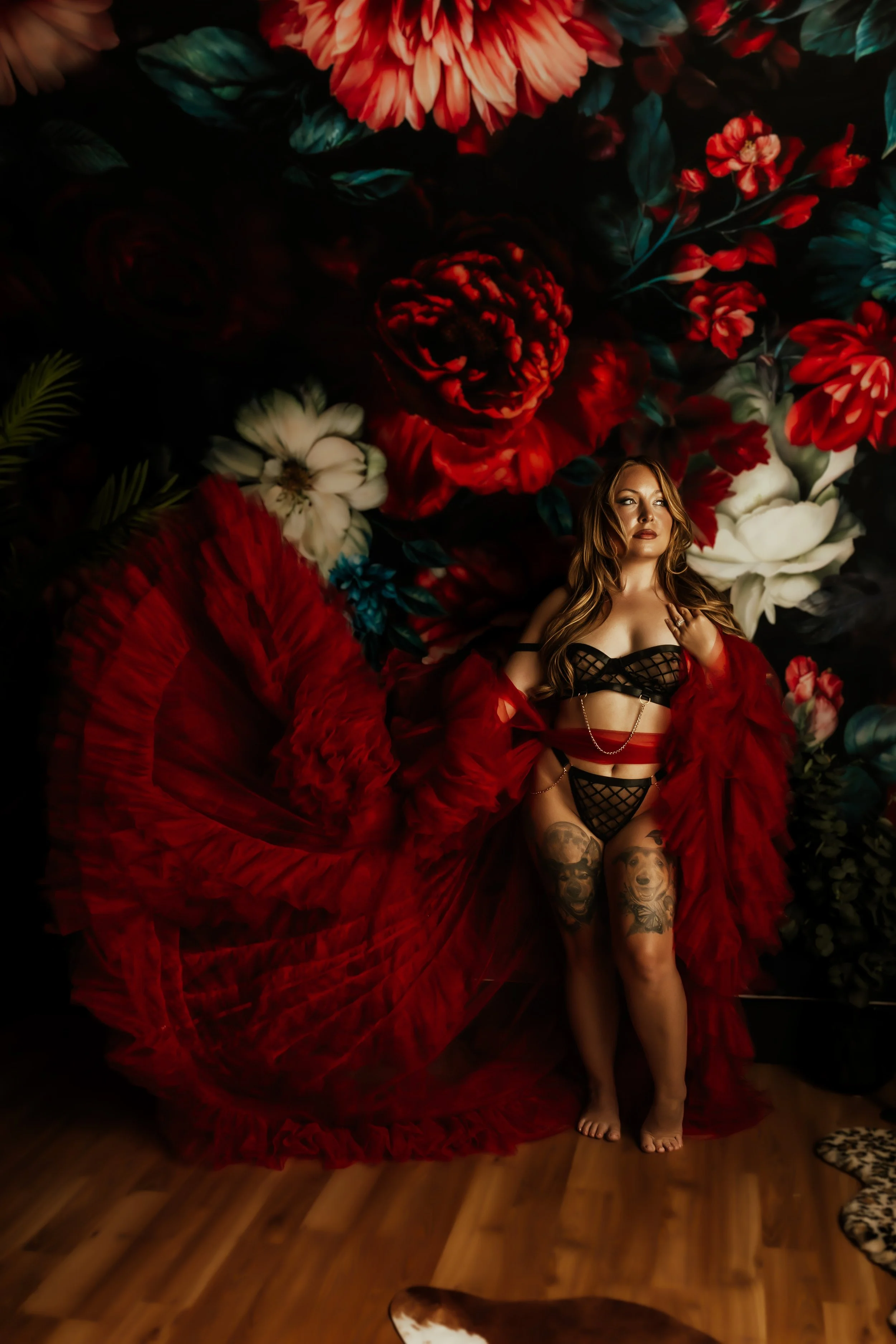 A woman with tattoos standing in front of a dark floral backdrop, wearing black lingerie and a red ruffled skirt or cape.