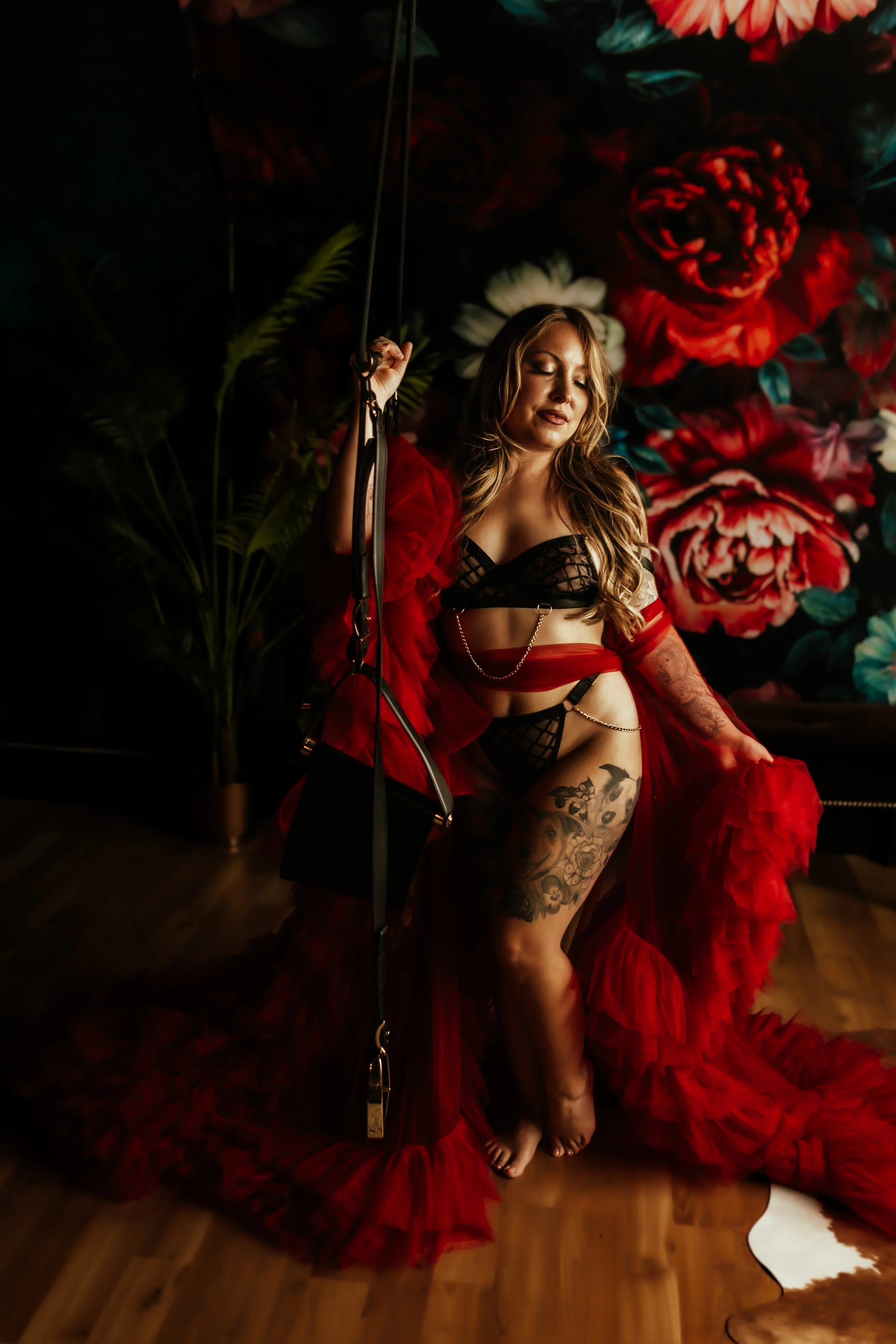 A woman with tattoos on her thigh and arm, wearing black lingerie and a red sheer robe, is sitting on a wooden floor with her eyes closed. She is holding onto a chain or strap, in front of a large floral mural backdrop.