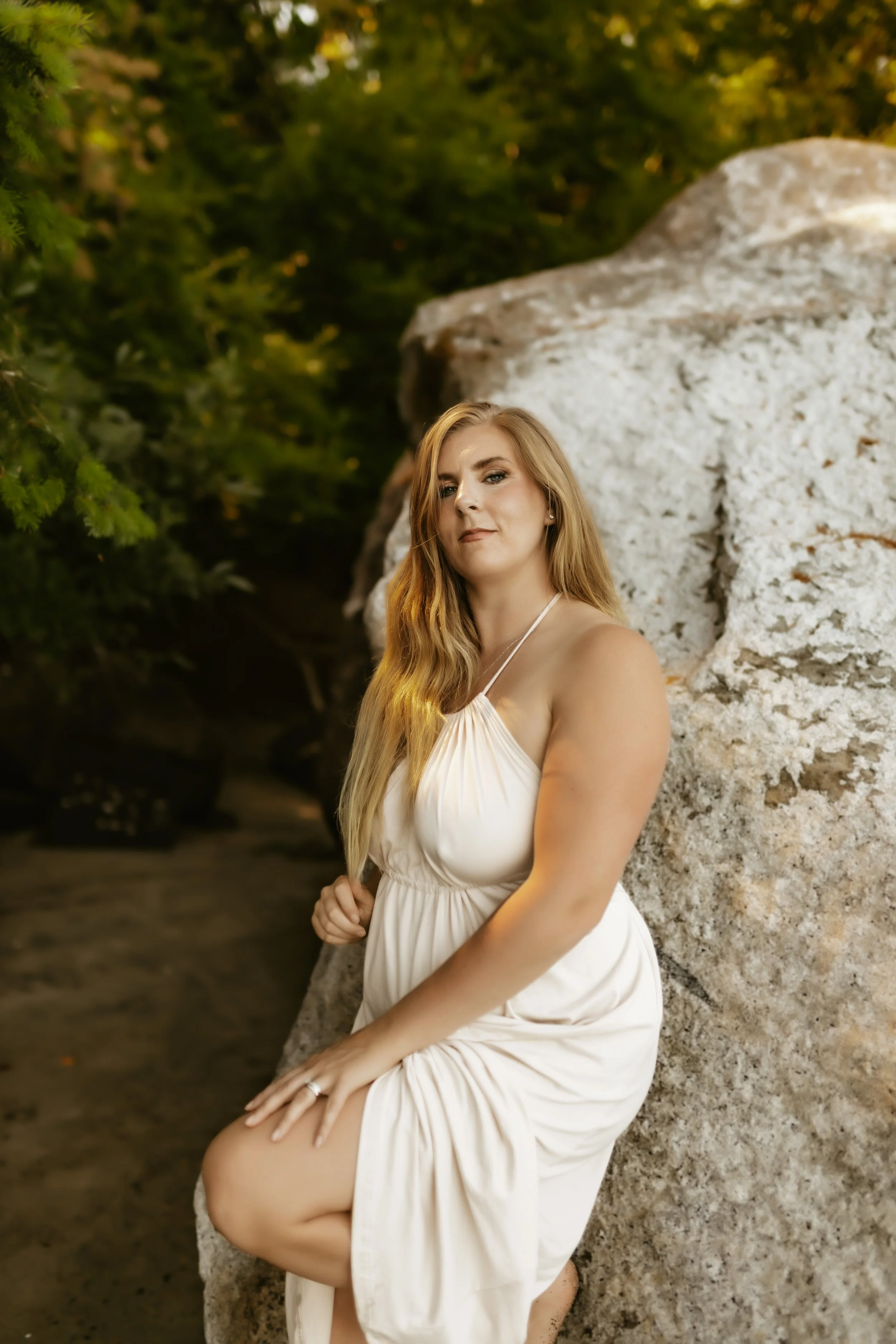 A blonde woman wearing a white dress sitting outdoors near a white rock, surrounded by green foliage.