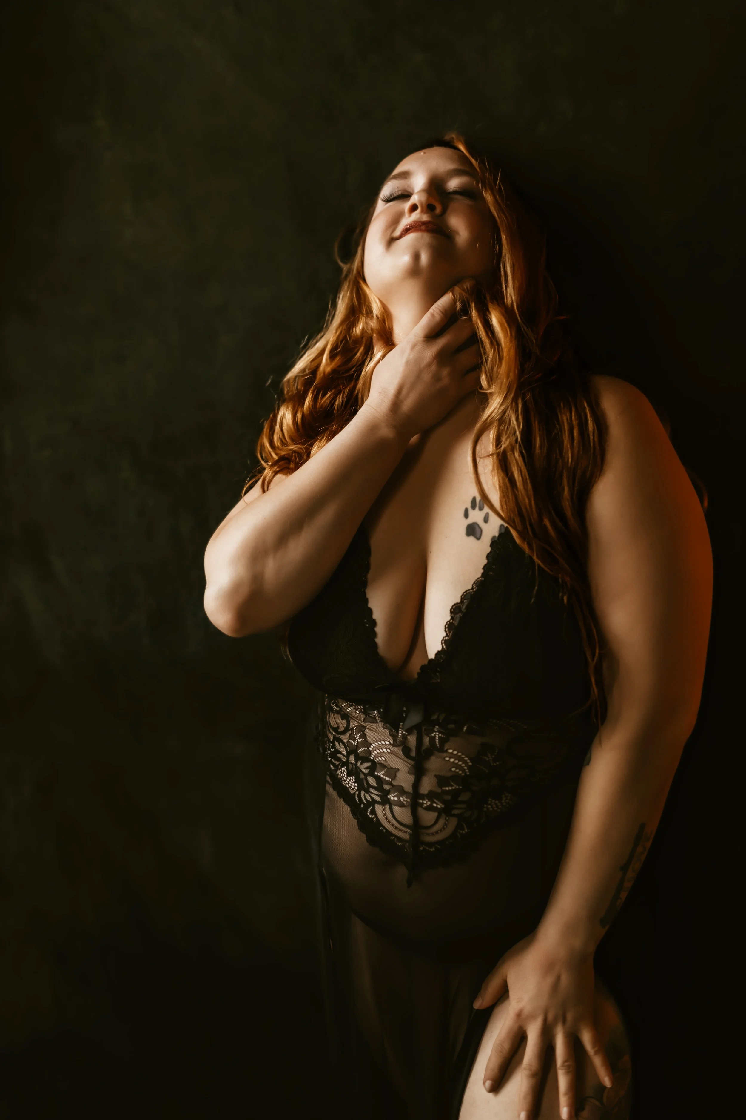 A woman with long red hair and a tattoo on her chest, wearing black lace lingerie, posing with her eyes closed and a hand gently touching her neck, in front of a dark background.