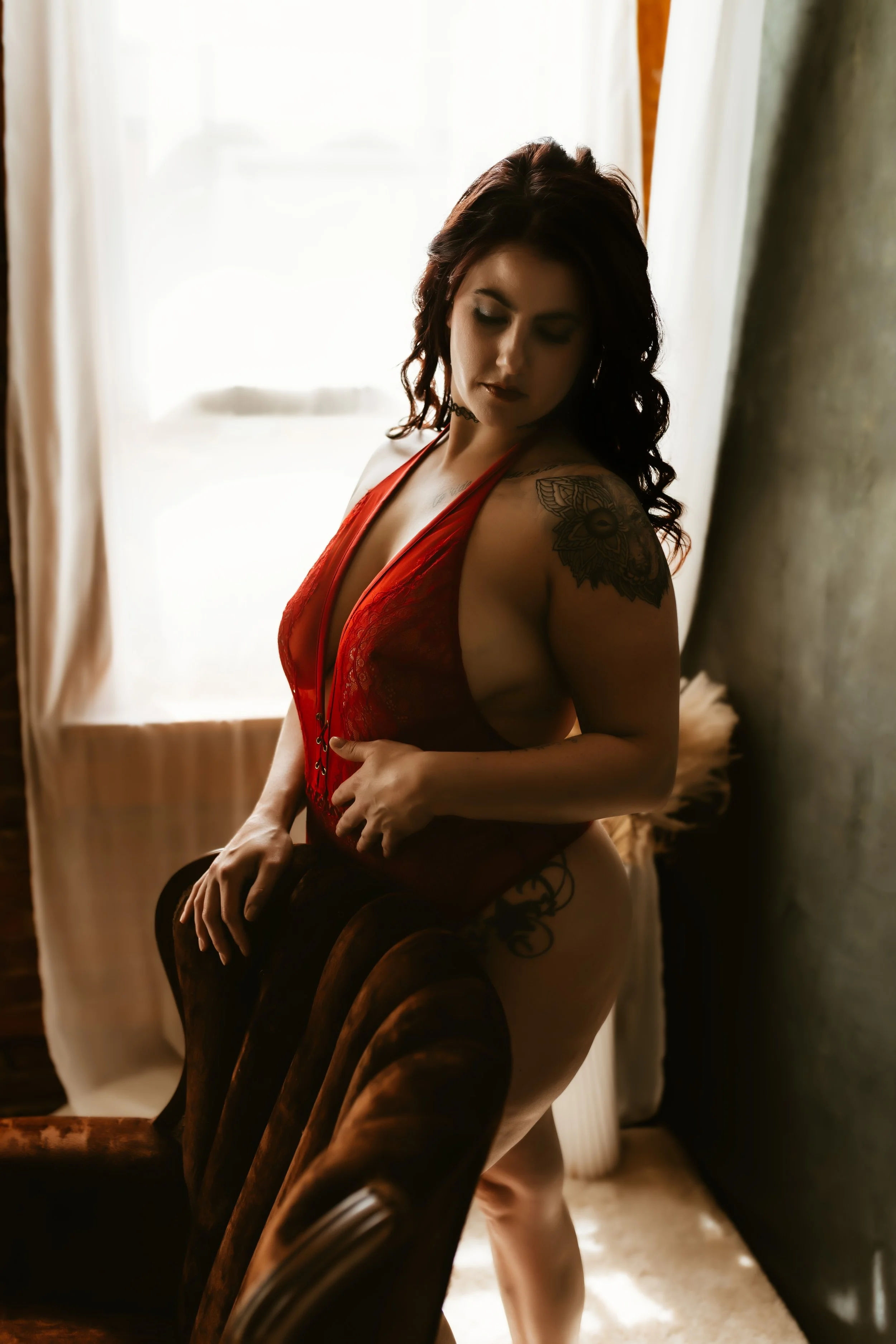 A woman with dark red hair, light skin, wearing a red lace bodysuit and black thigh-high stockings, sitting on a vintage wooden chair near a window with sheer curtains and a curtain on the side, in soft natural lighting.