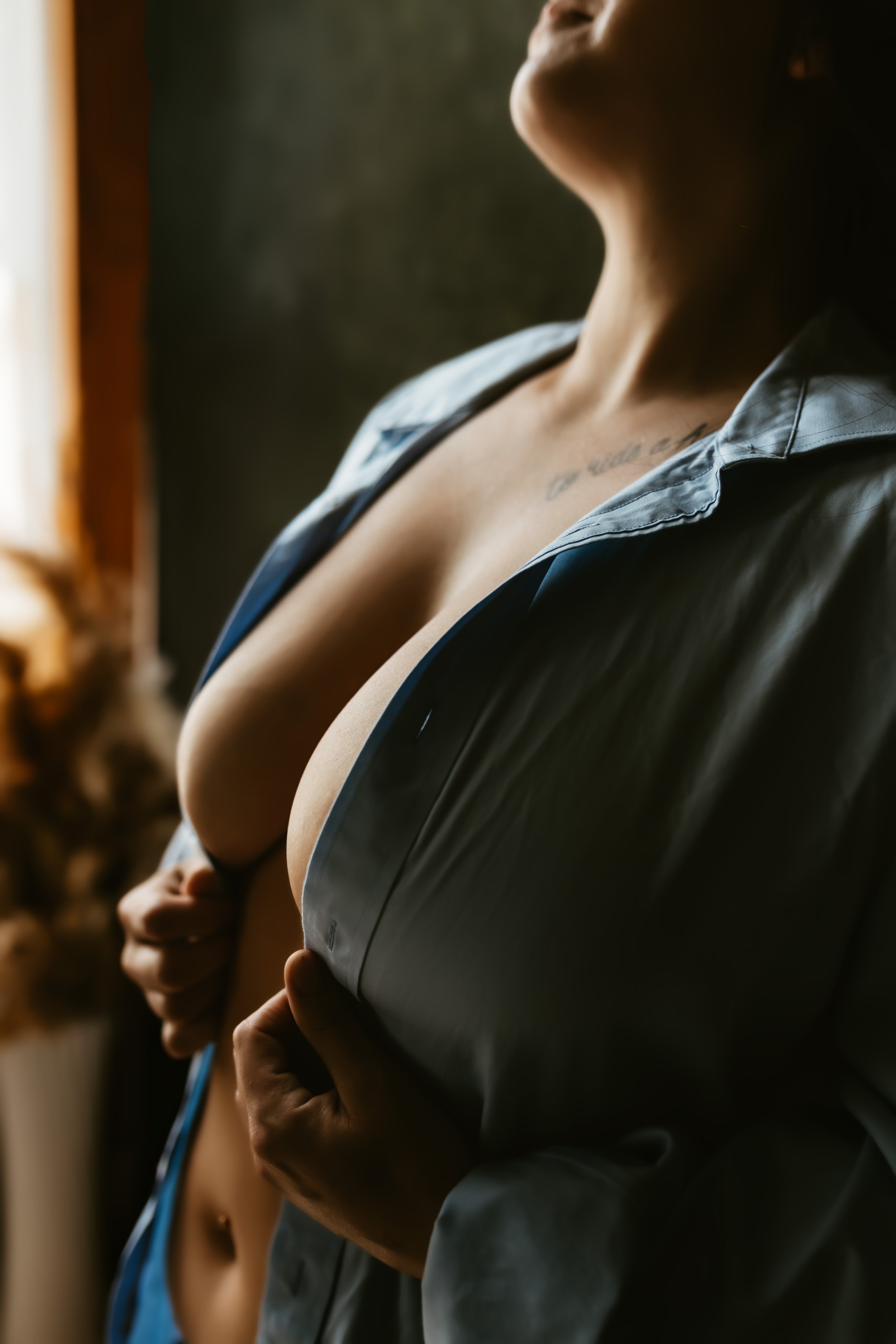 A woman with a tattoo on her chest is standing near a window, with a person holding a stethoscope behind her, suggesting a medical examination.