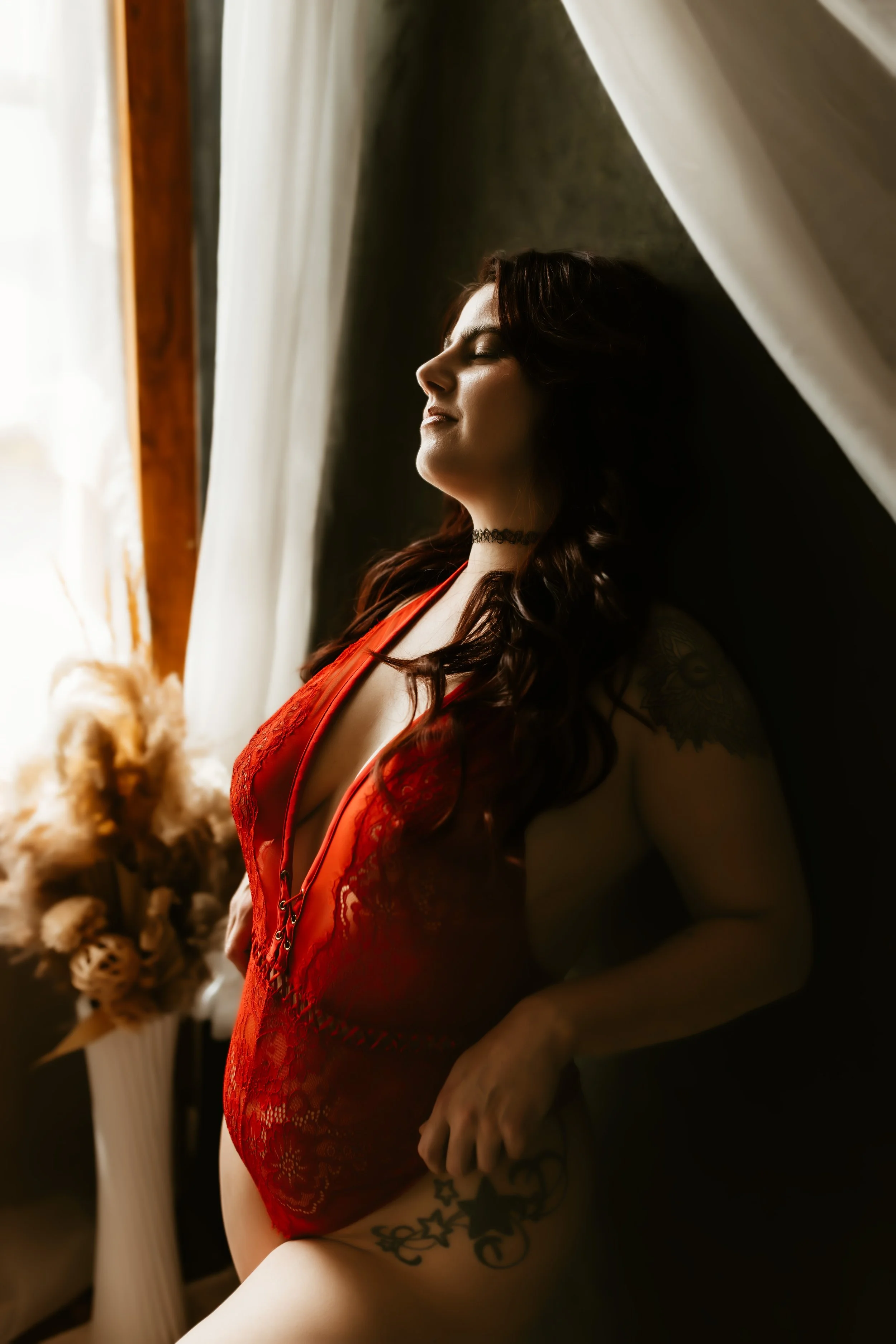A woman with dark wavy hair and tattoos wearing a red lace lingerie, standing by a window with closed white curtains, relaxing with eyes closed.