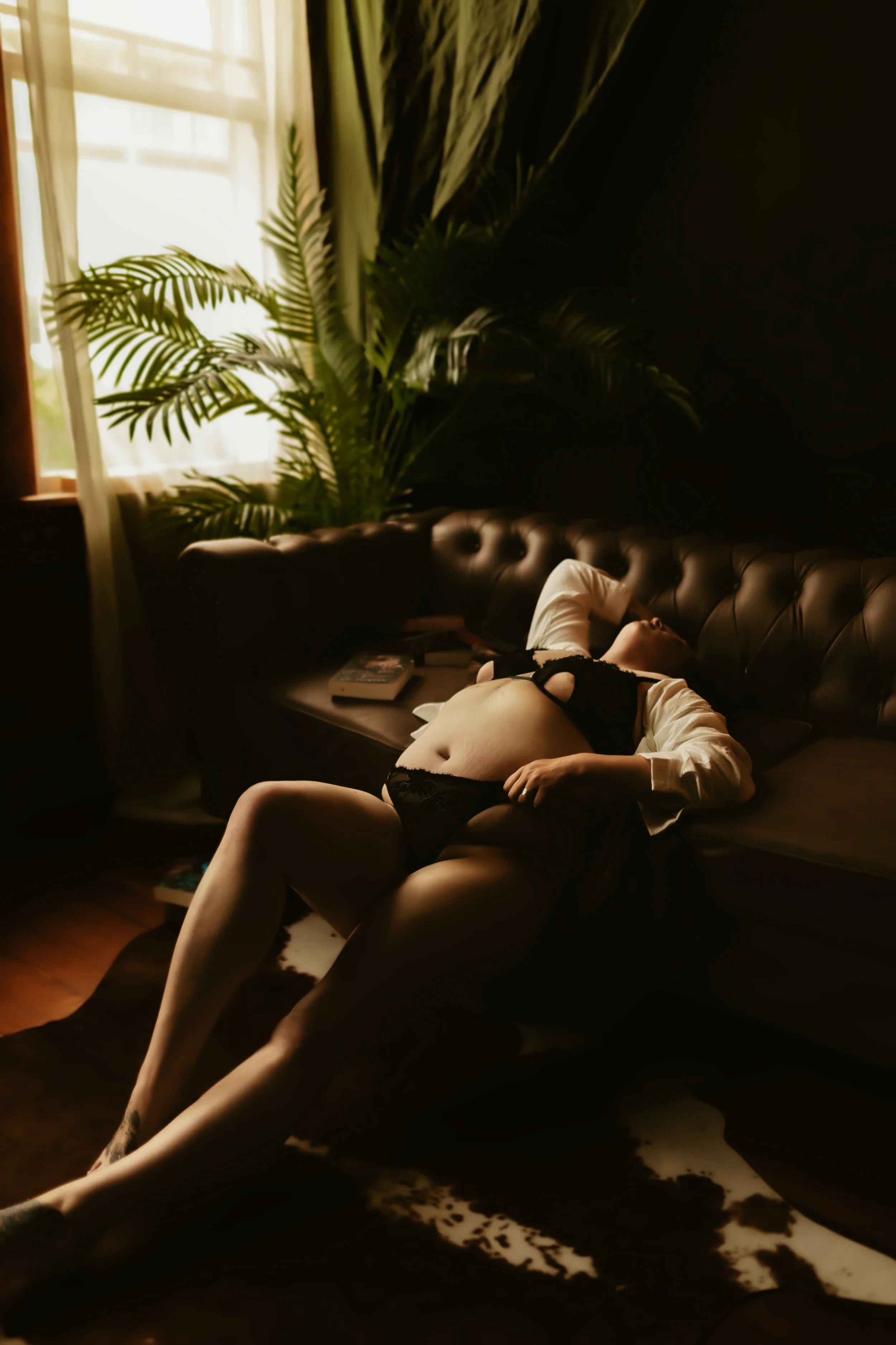 A woman lying on a dark sofa in a room with a large potted plant, curtains, and sunlight coming through the window.