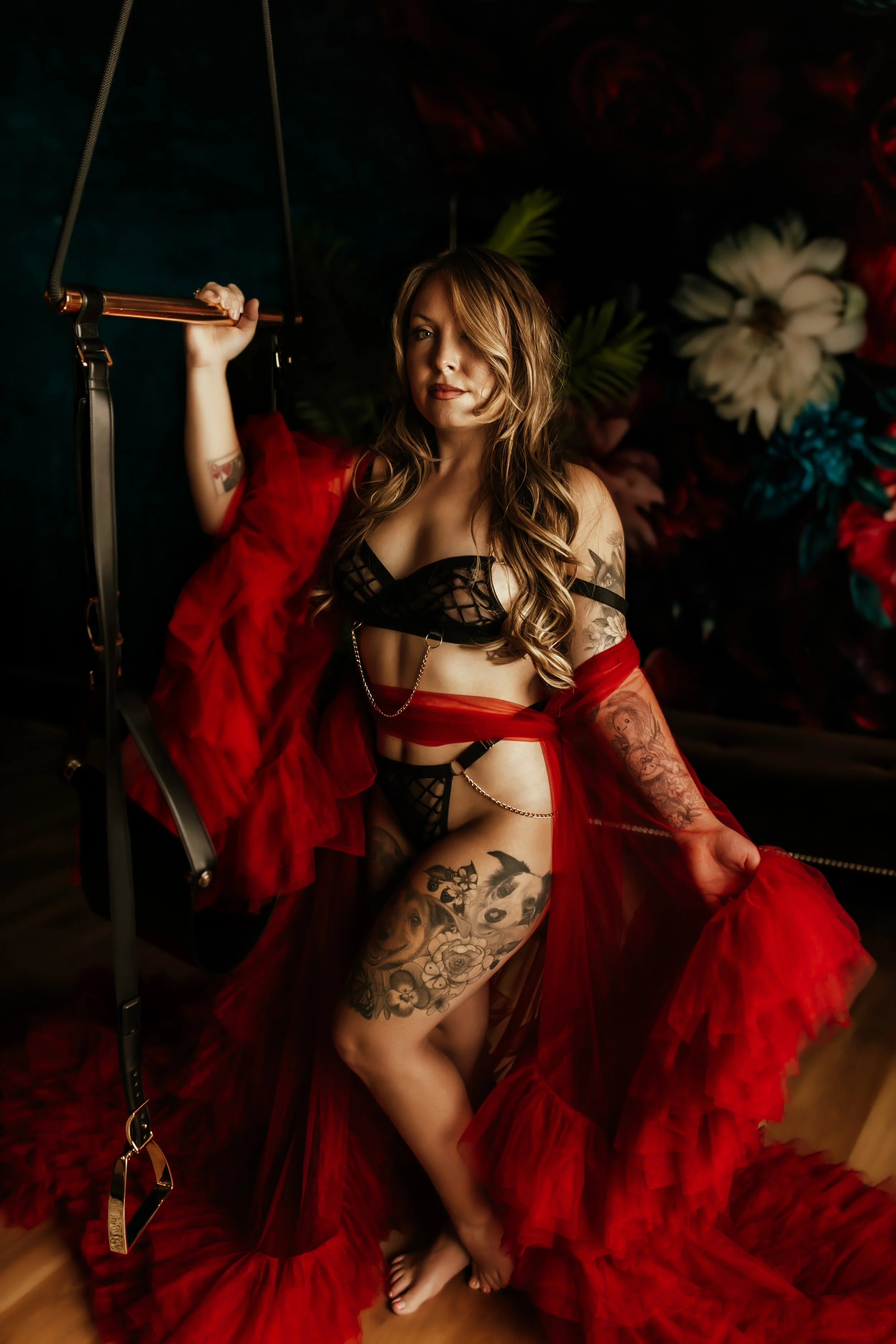 A woman with long wavy hair and tattoos on her leg and arm is sitting on a red velvet fabric in front of a dark floral background. She wears black lingerie and a red sheer robe, holding a horizontal bar with one hand. She has a confident expression a