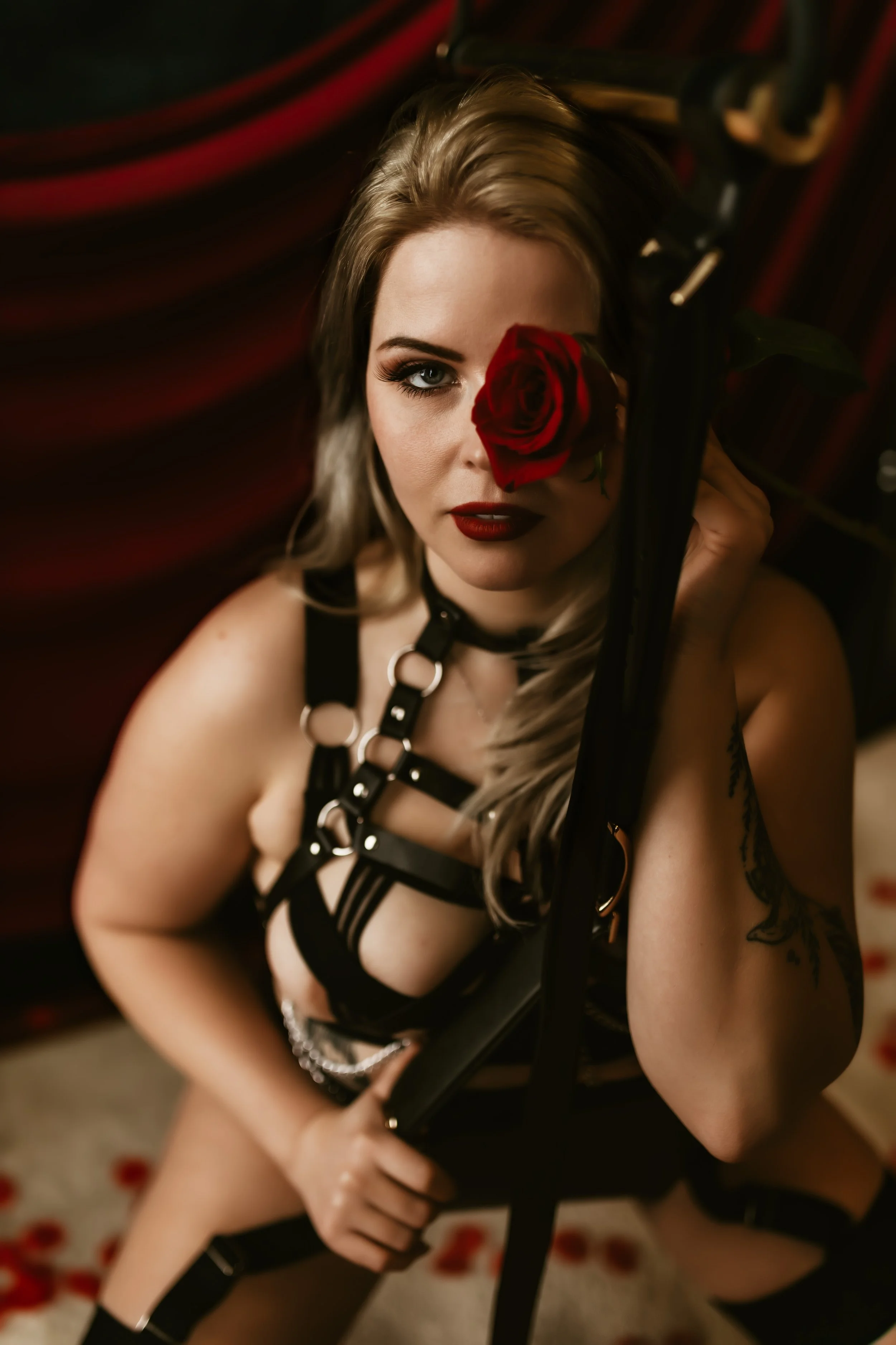 A woman with blonde hair and makeup holding a red rose over one eye, wearing black lingerie with straps, in a dark setting with a blurred red and black background.