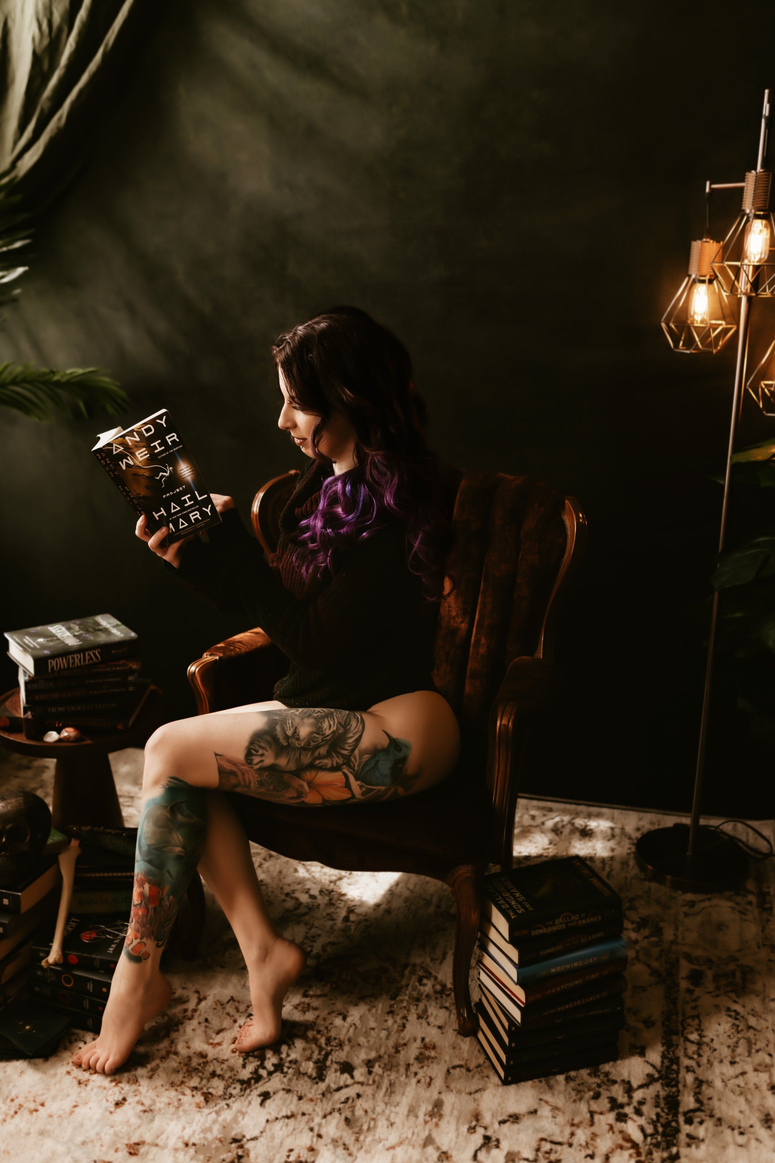 A woman with purple hair, tattoos on her legs, and a black sweater sitting in a vintage armchair in a dimly lit room, reading a book, with several stacks of books around her, decorative lighting, and plants.
