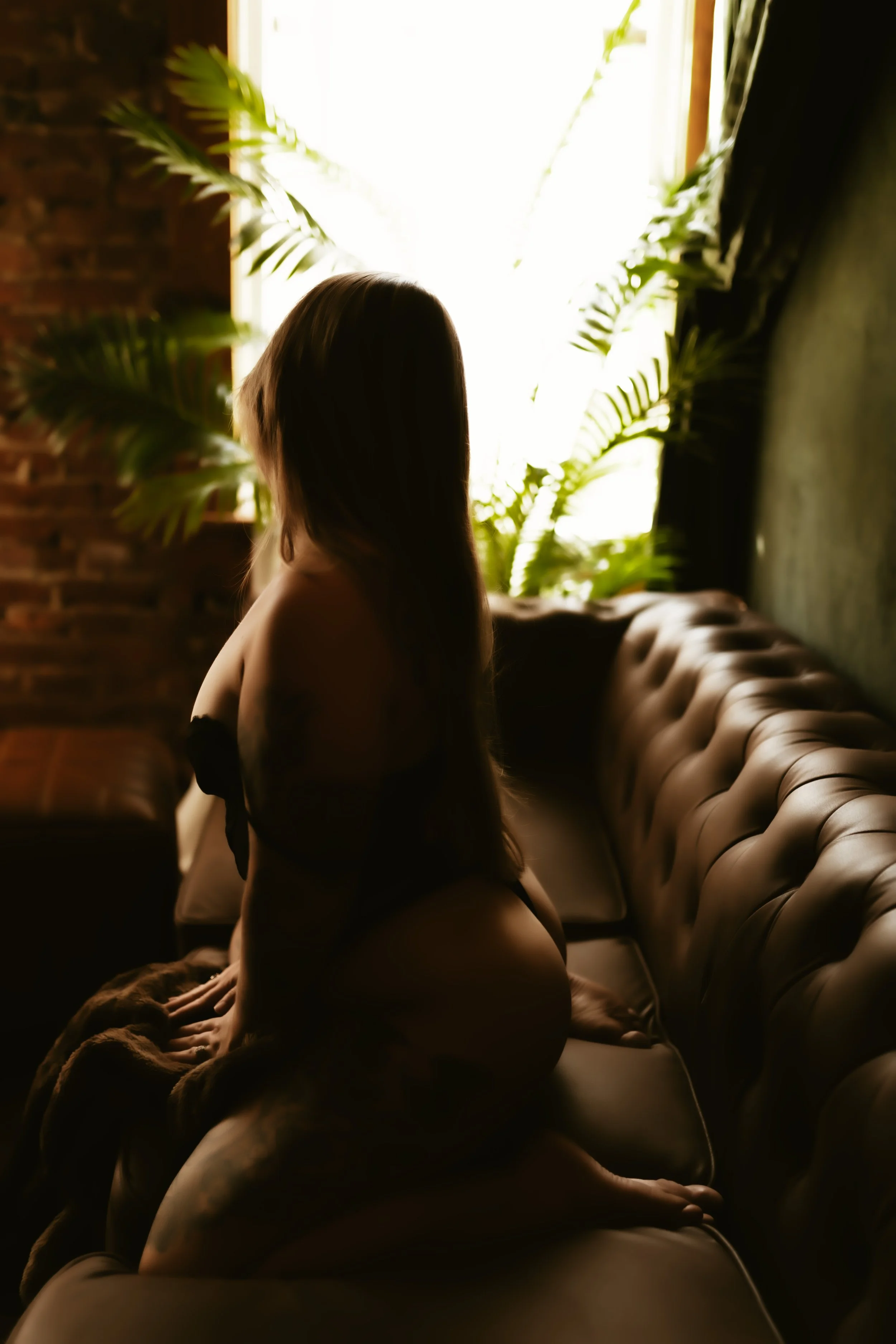 A woman with long hair sitting on a beige tufted sofa near a window with bright light, surrounded by large green plants in a cozy, dimly lit room.