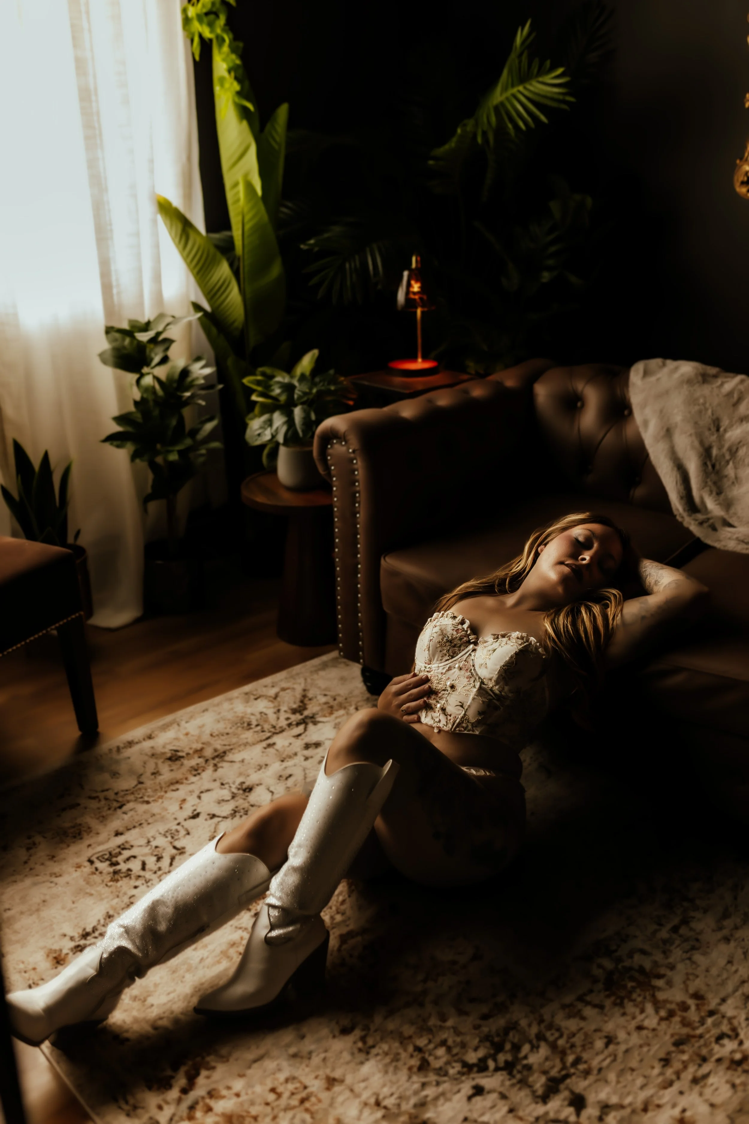 A woman with long blonde hair lying on a patterned rug on the floor, wearing a floral top and sparkly white thigh-high boots, with her eyes closed and arm resting on her head. The room has a black wall with lush green plants, a leather sofa with a li