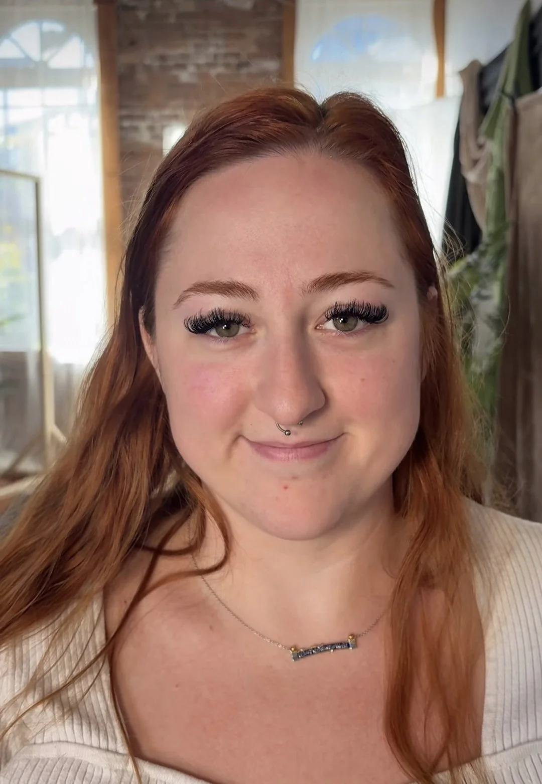 A woman with red hair and piercings, wearing a white top and necklace, smiling indoors with natural light coming through windows.