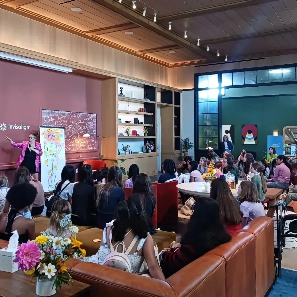 A woman in a pink jacket speaks to an audience in a modern, well-lit room with a high ceiling, wooden paneling, and large windows. The audience is seated on couches and chairs, attentively listening. There are colorful decorations, flowers, and artwork in the background.
