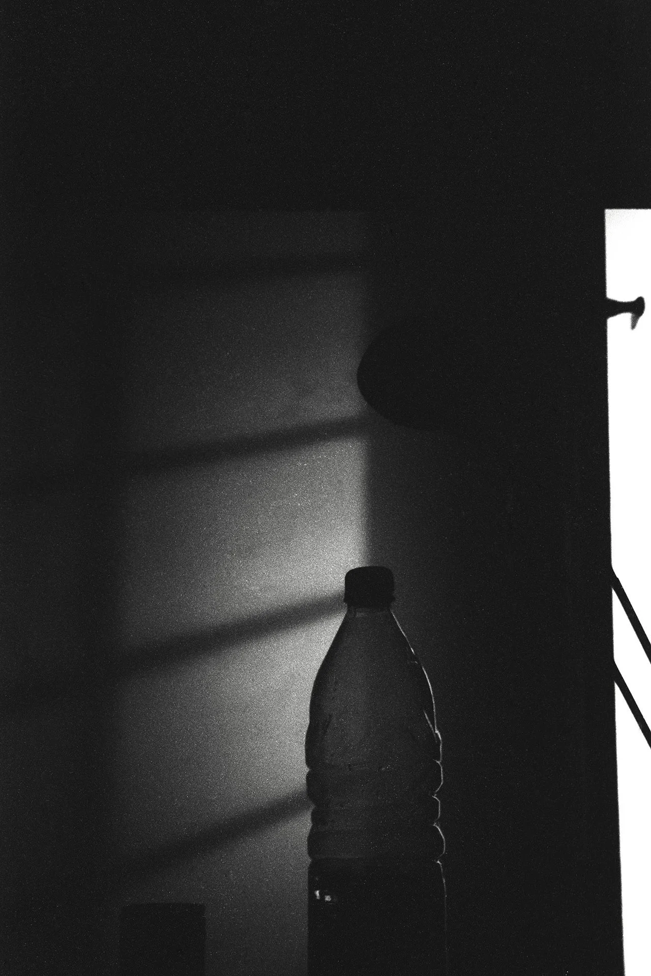 Black and white photo of a plastic water bottle with a label on it, placed on a surface near a wall or flat surface with shadows cast on the wall.